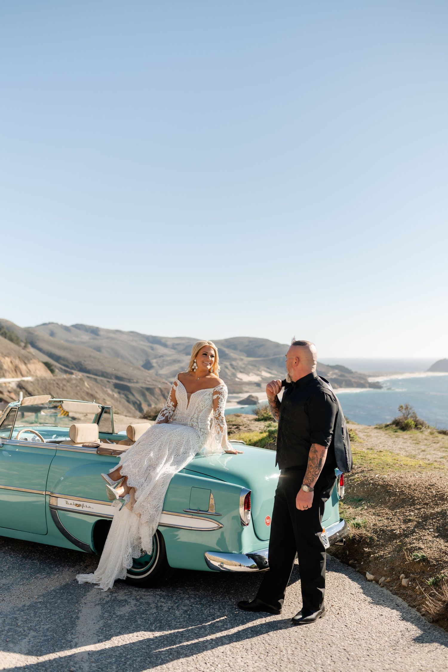 Scenic coastal wedding photoshoot with classic turquoise car against mountain backdrop.