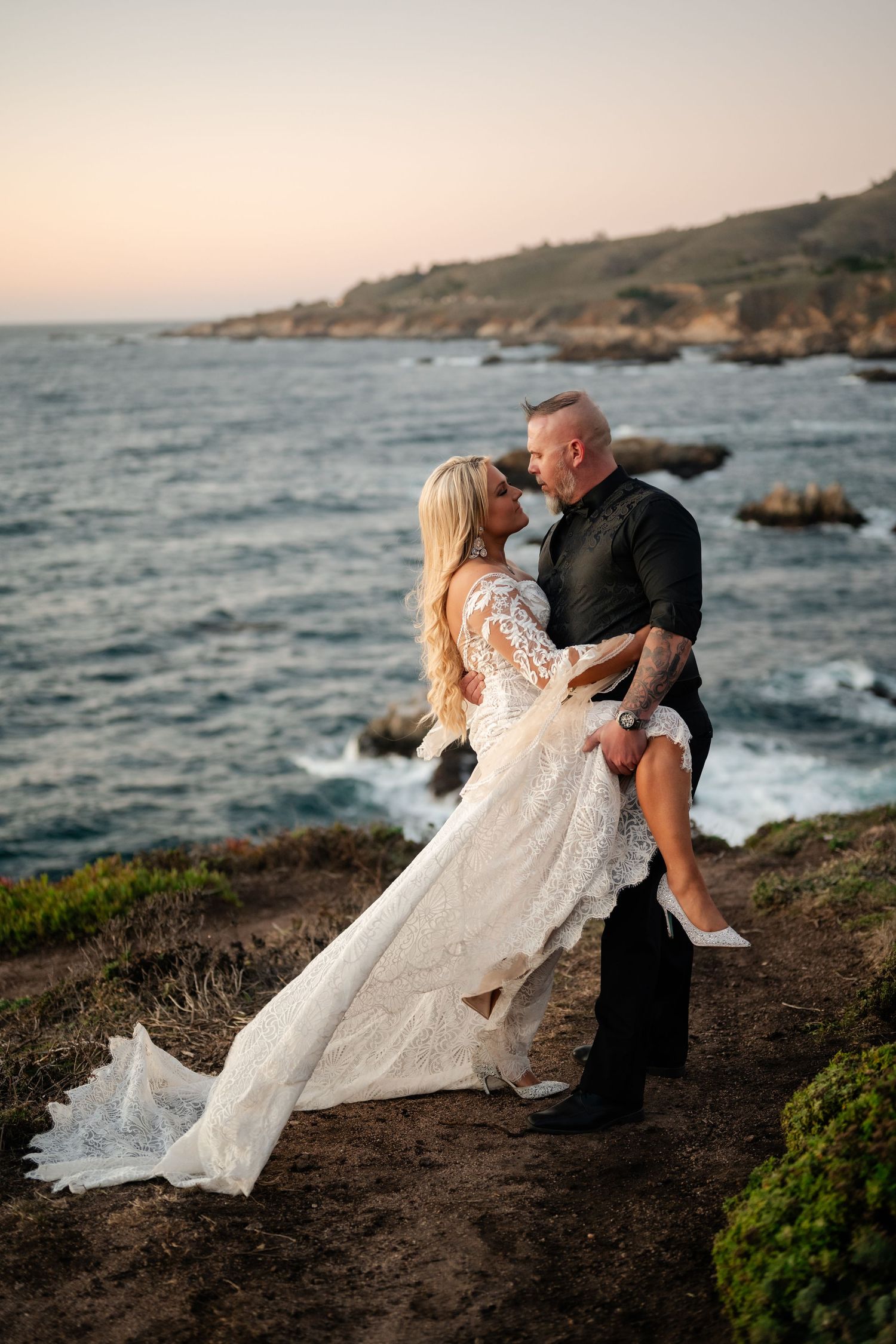A romantic wedding photo on a coastal cliff with crashing waves and sunset in the background.