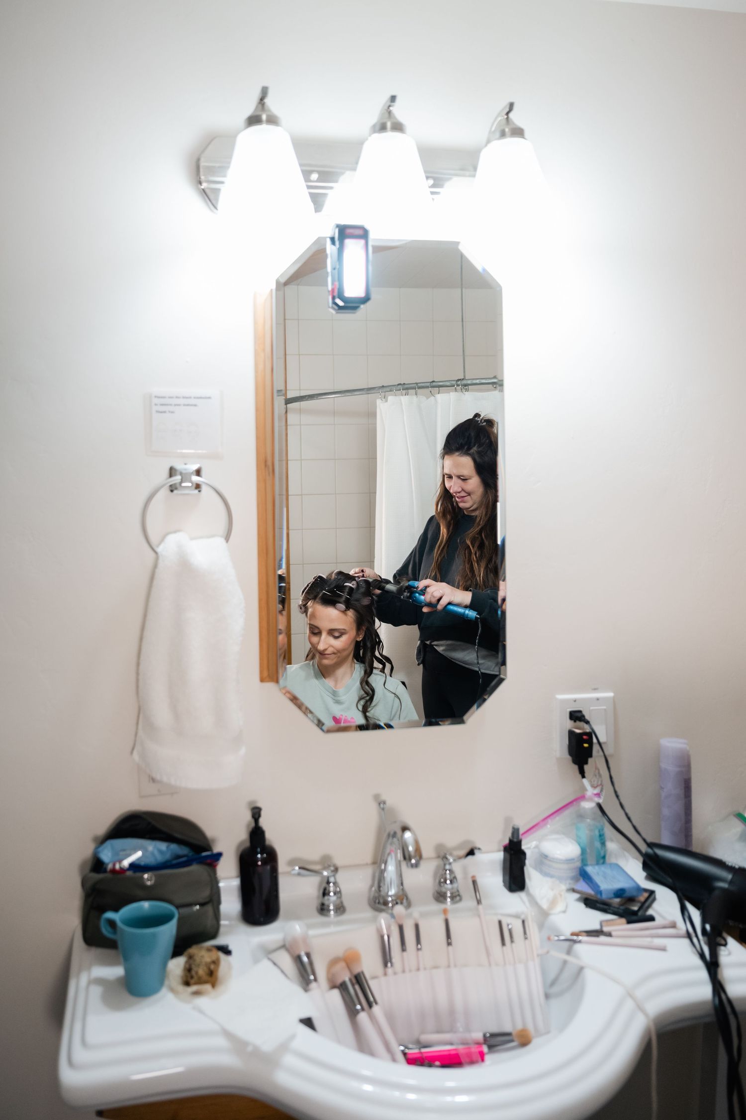 Hair styling session reflected in bathroom mirror with professional tools and equipment on white countertop.