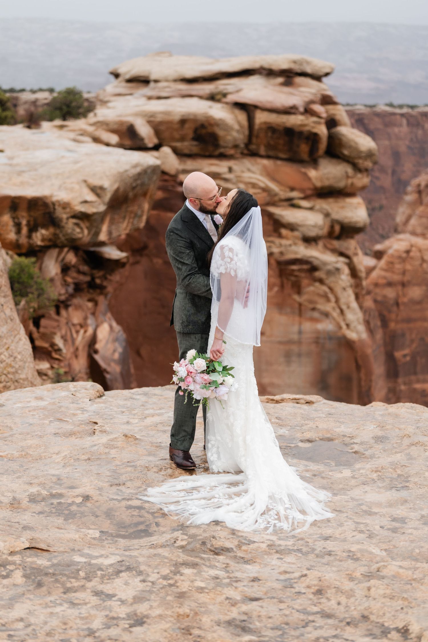 A wedding couple embraces on the edge of a dramatic desert canyon overlook.