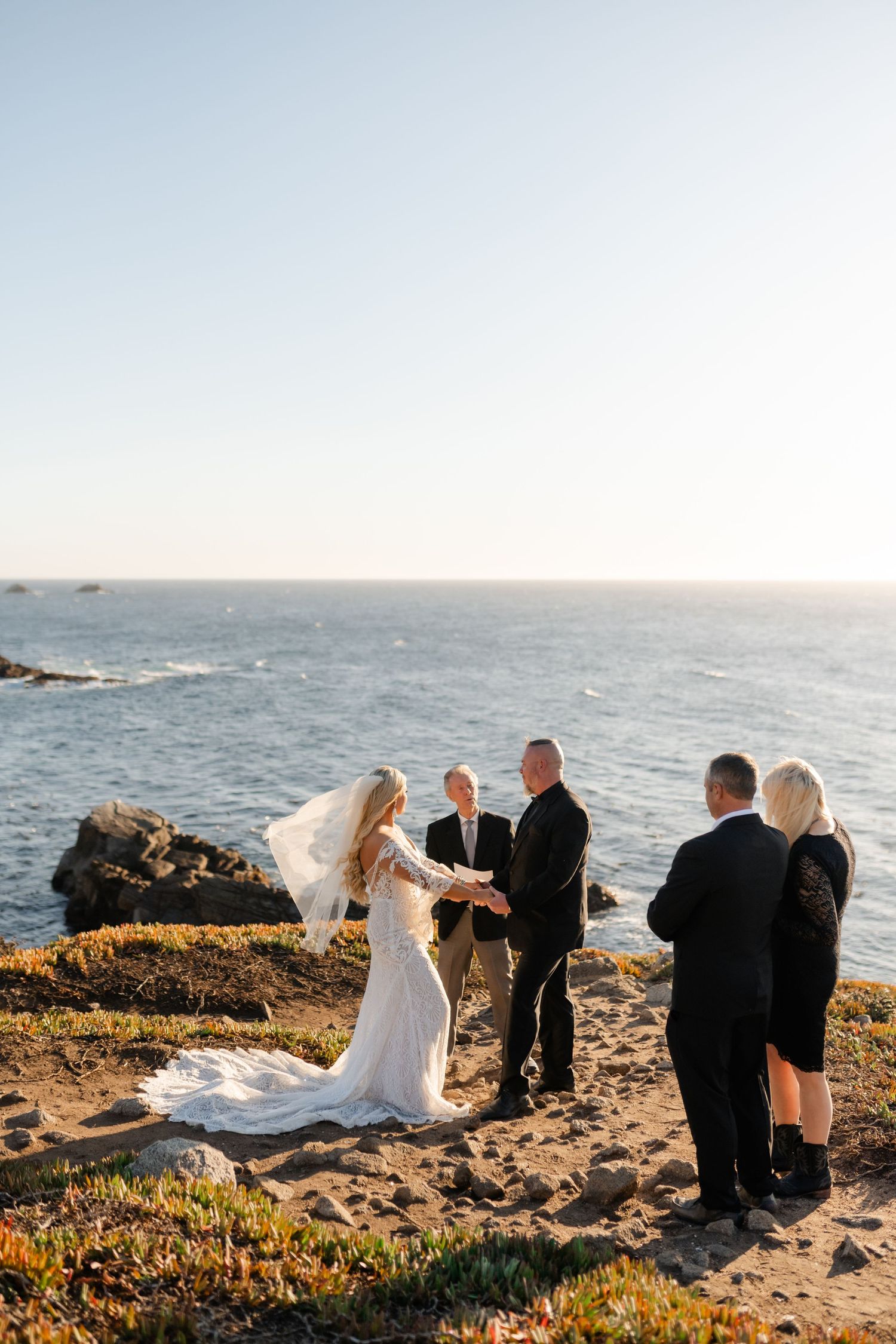 A small wedding ceremony takes place on a scenic coastal bluff during golden hour.