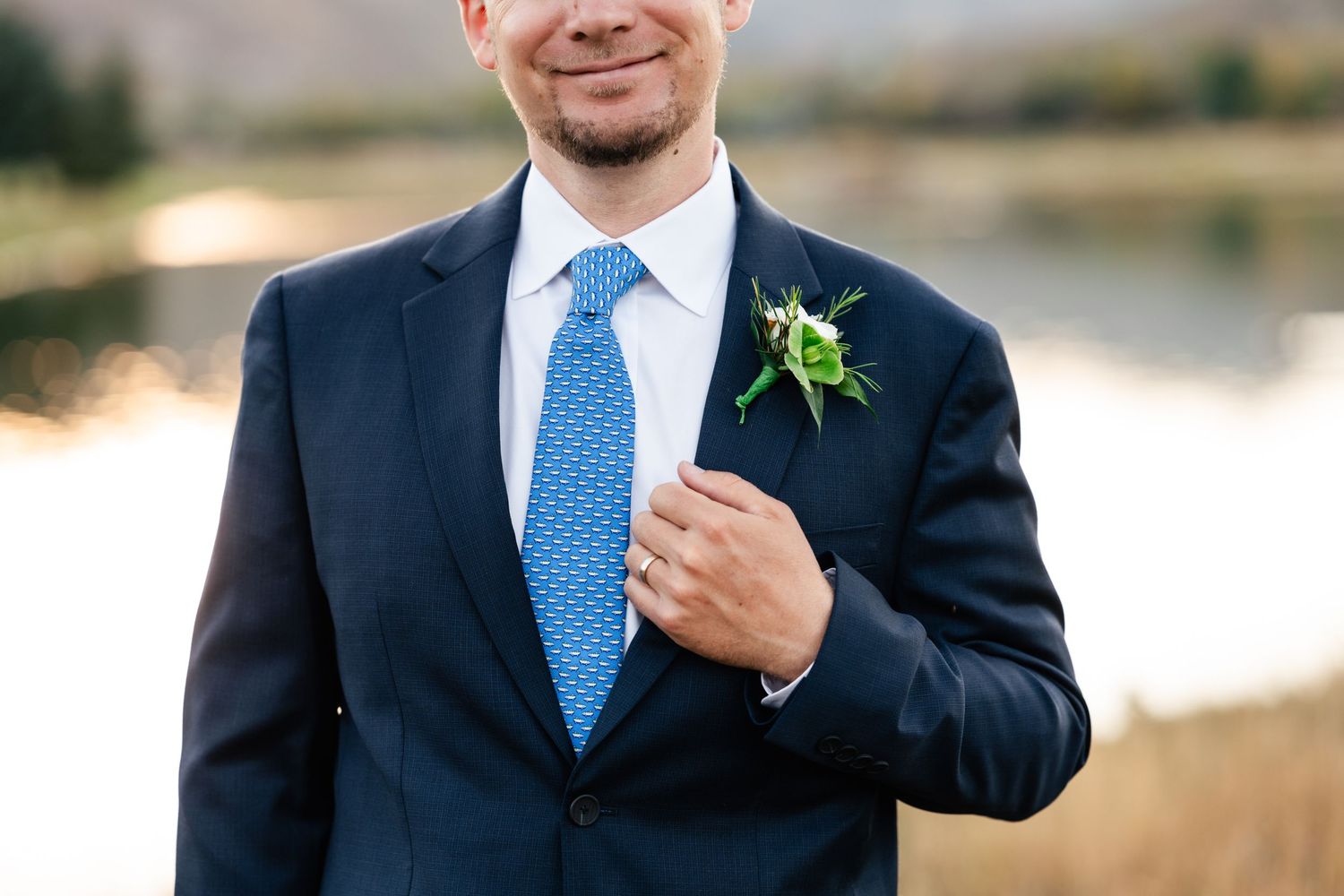 Groom in navy suit with blue tie and boutonniere smiles during outdoor wedding portraits.