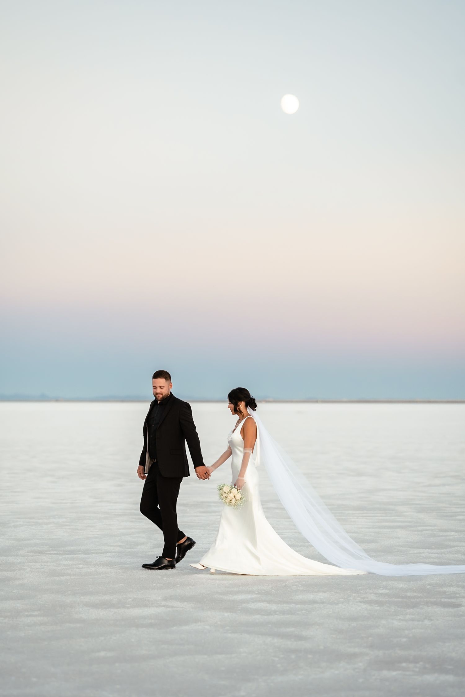 A couple in wedding attire walks along a serene salt flat at sunset with a full moon rising in the pastel sky. Salt Flats Photographer.