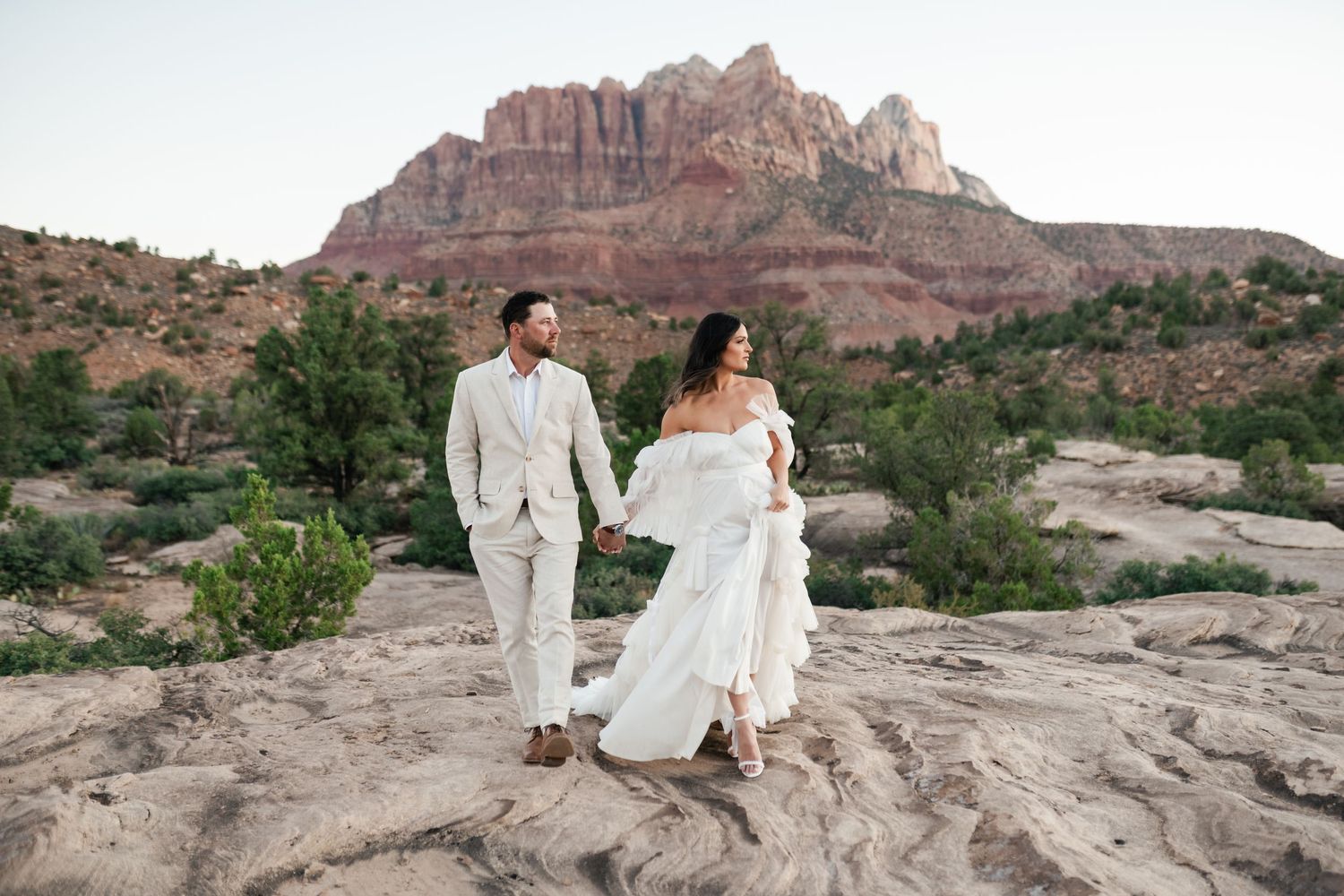 A couple in wedding attire stands on red rock formations in the desert landscape during sunset.