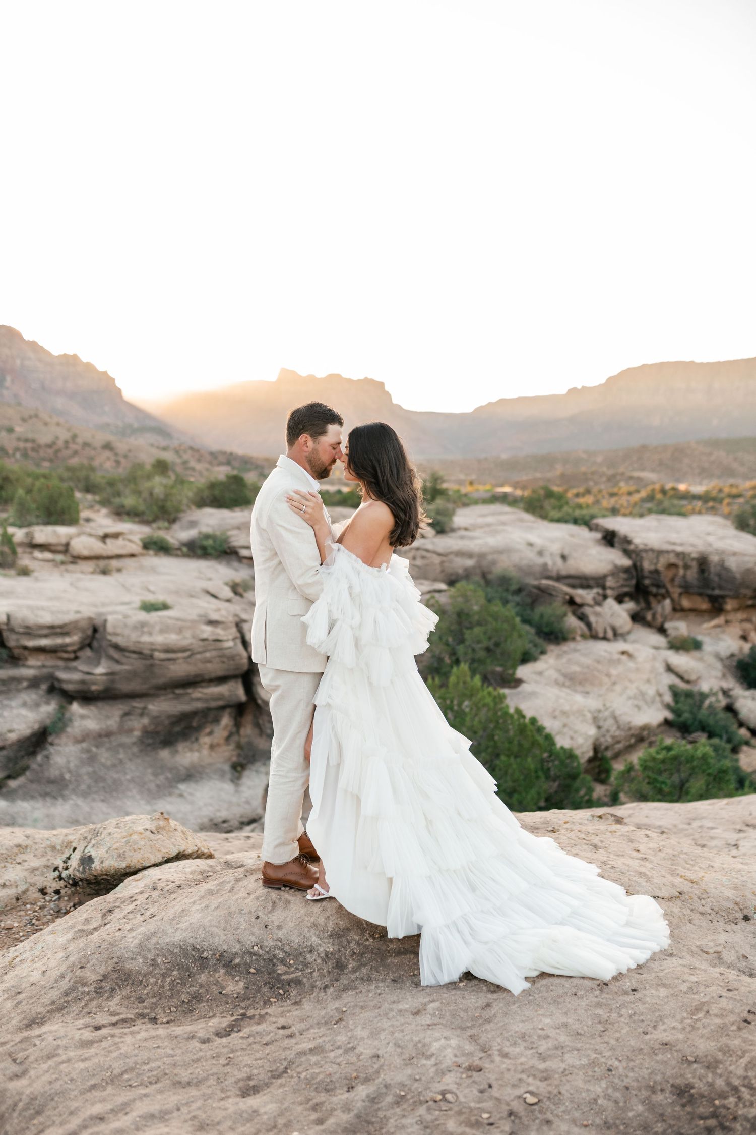 Zion Adventure Session. A couple in wedding attire embrace on a rocky cliff overlooking a scenic desert canyon landscape at sunset.