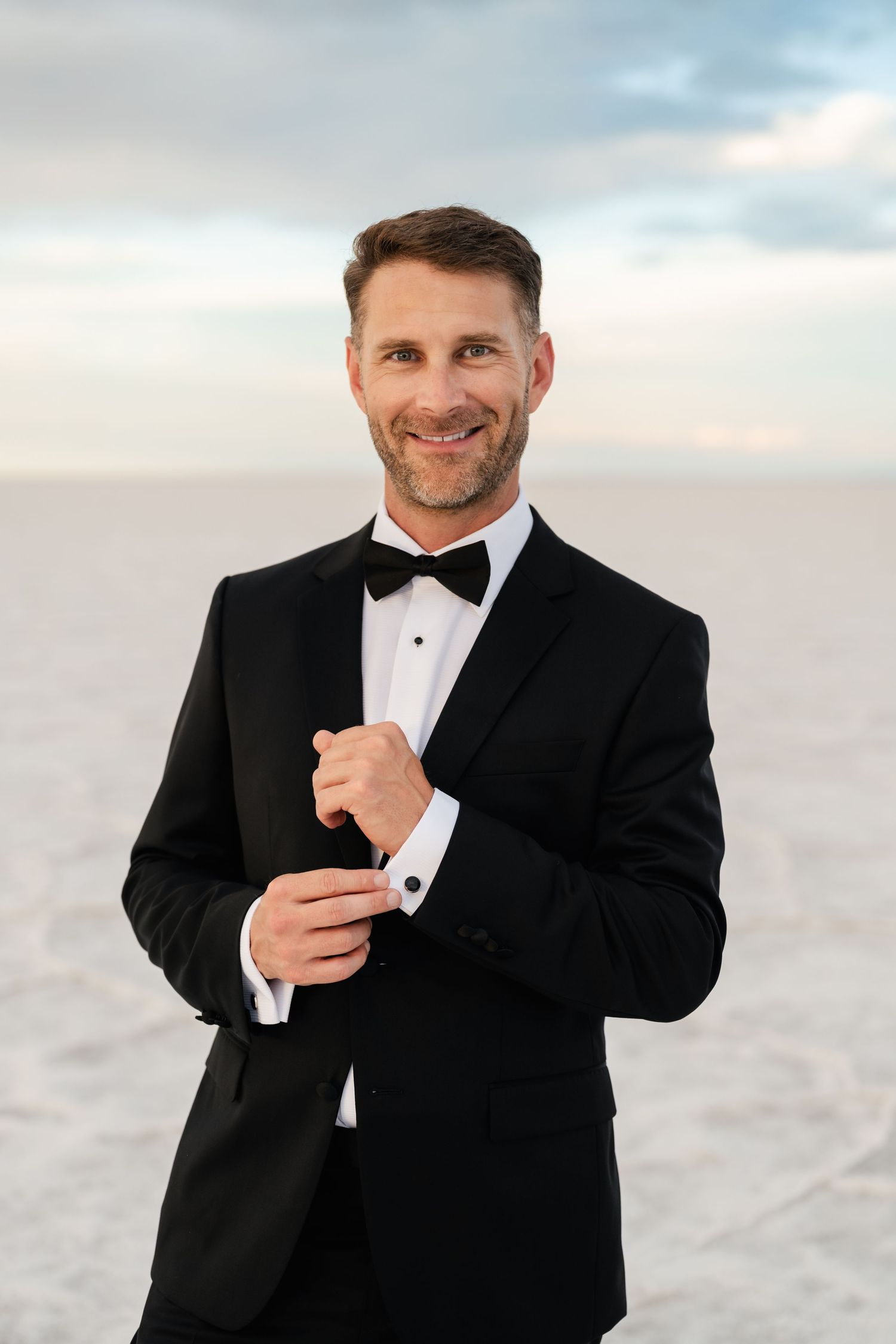 Salt Flats Elopement Photographer. A groom in a classic black tuxedo and bow tie adjusts his cuff against a cloudy sky backdrop.