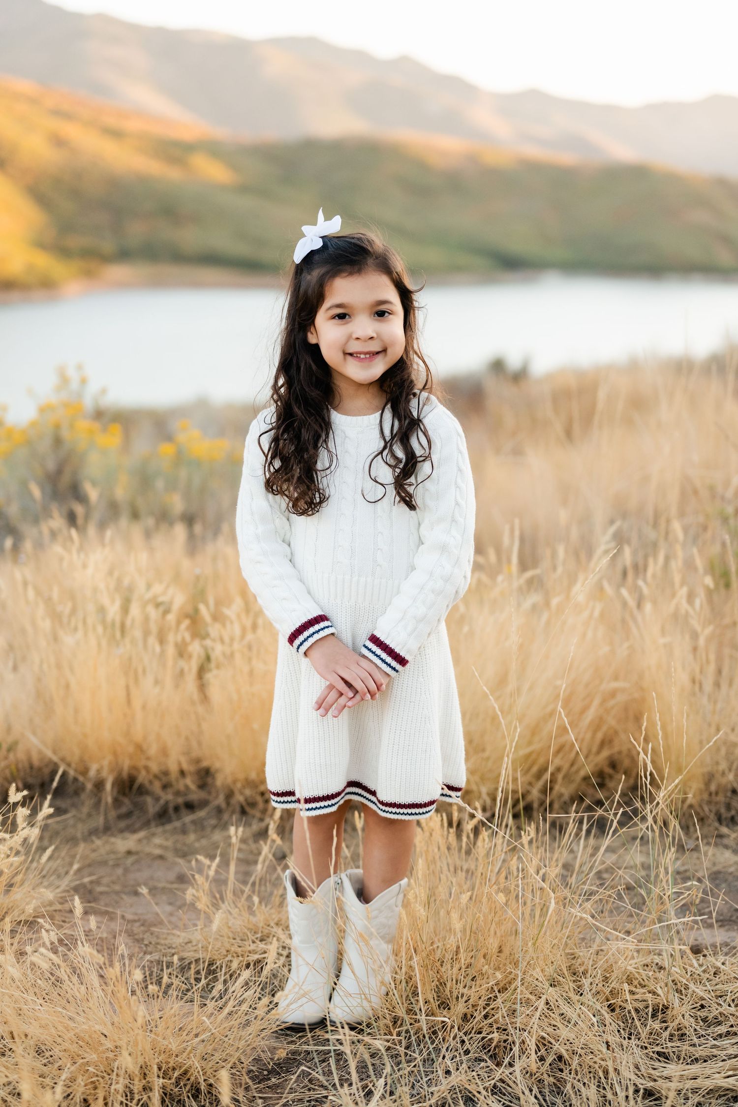 A child in a white sweater dress and white boots stands in golden grass near a lake with mountains in the background.