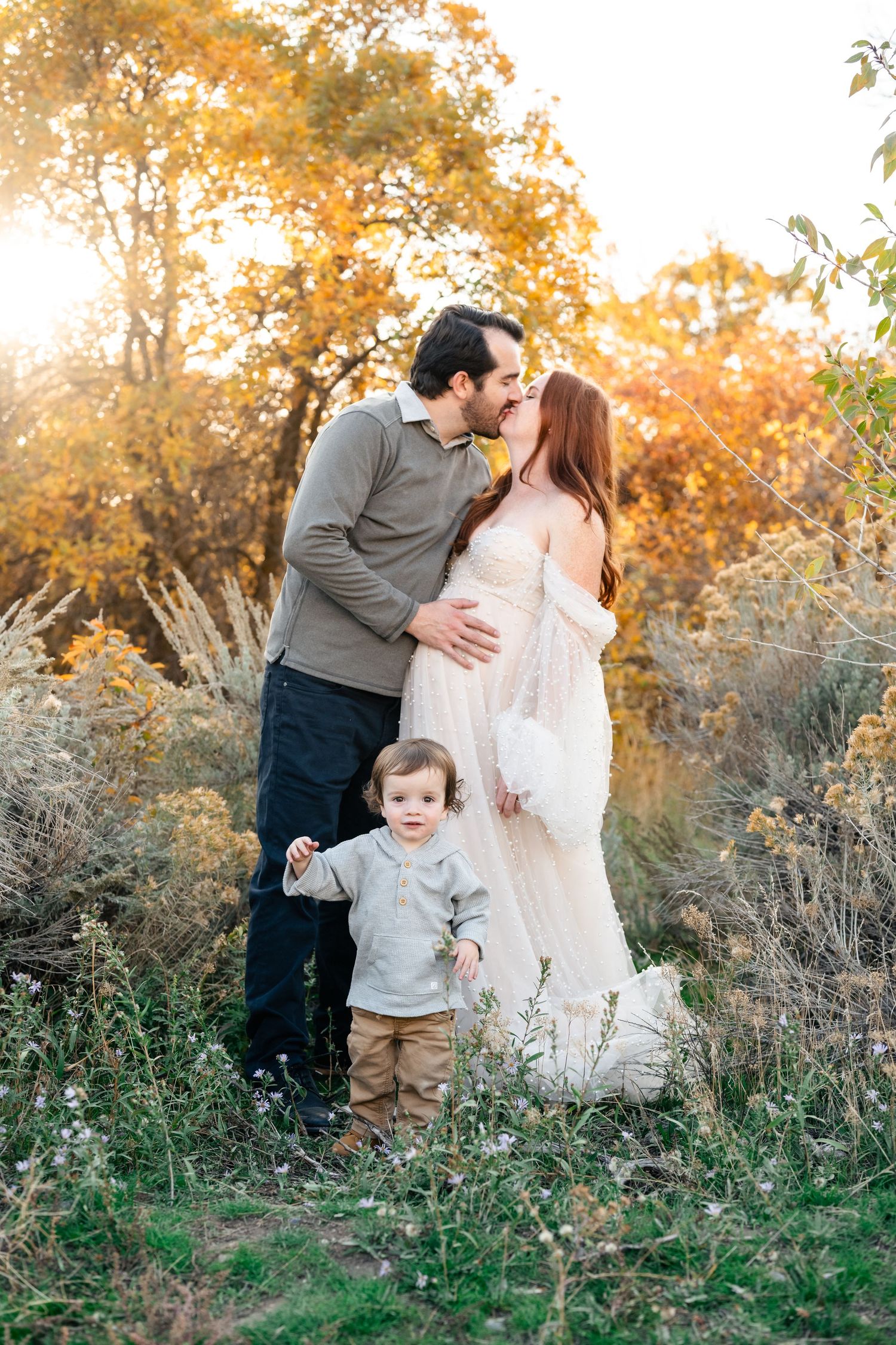 Family shares tender moments together in a golden autumn field during sunset with tall grass and foliage.