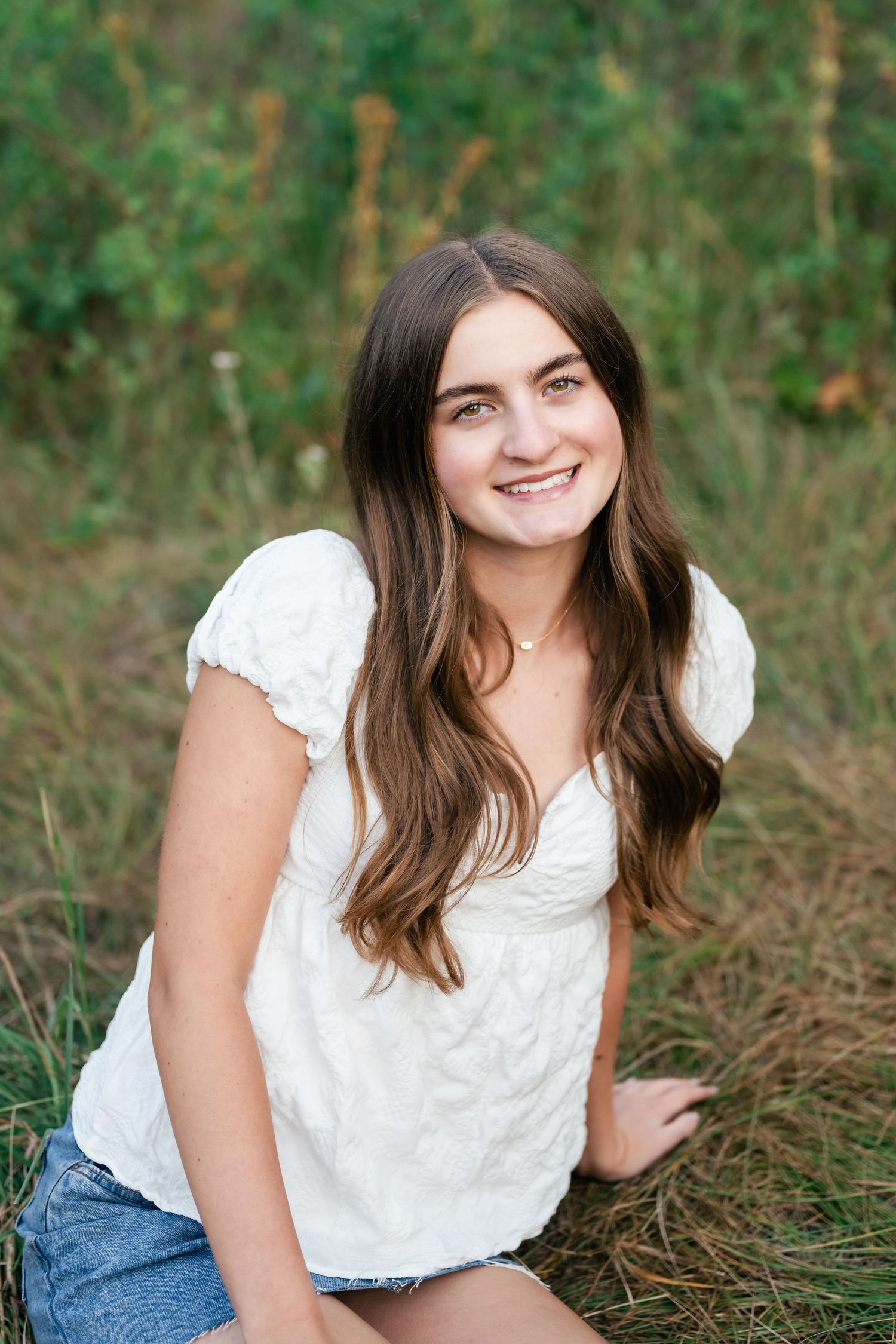 A person in a white top and jeans sits in a grassy outdoor setting with natural lighting and a candid pose.