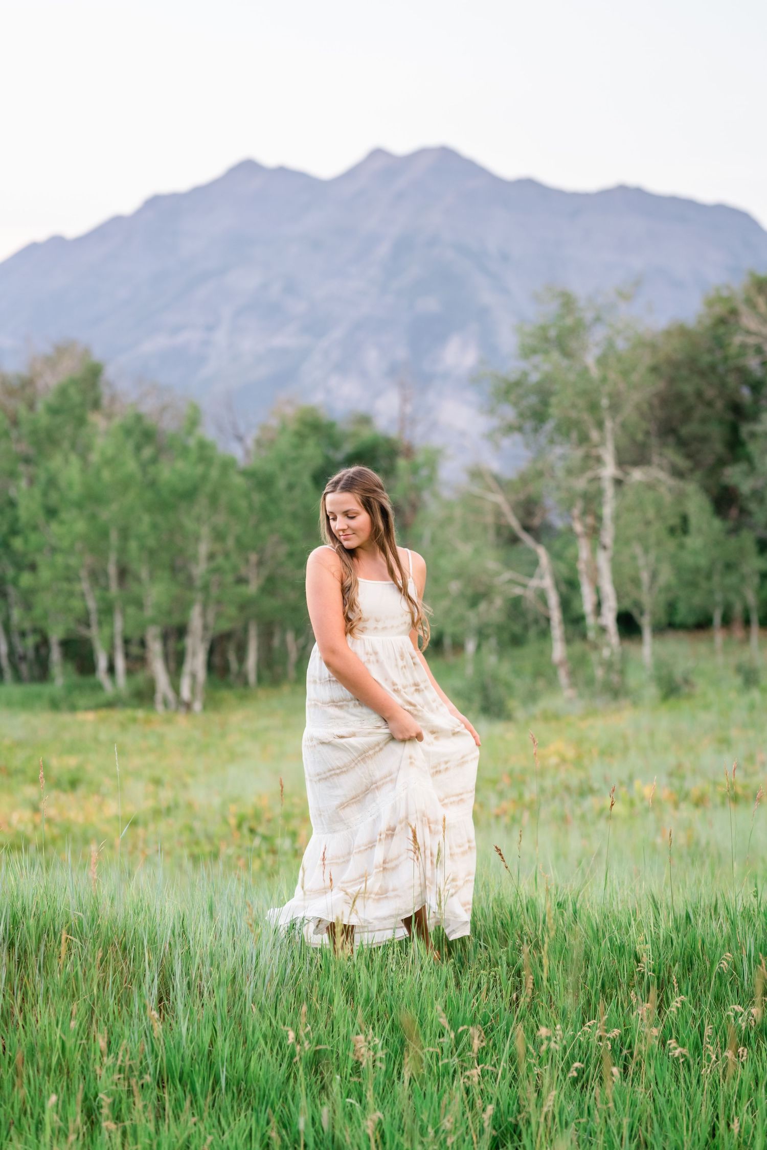 A woman in a white dress sits peacefully in a mountain meadow surrounded by wildflowers and aspen trees.