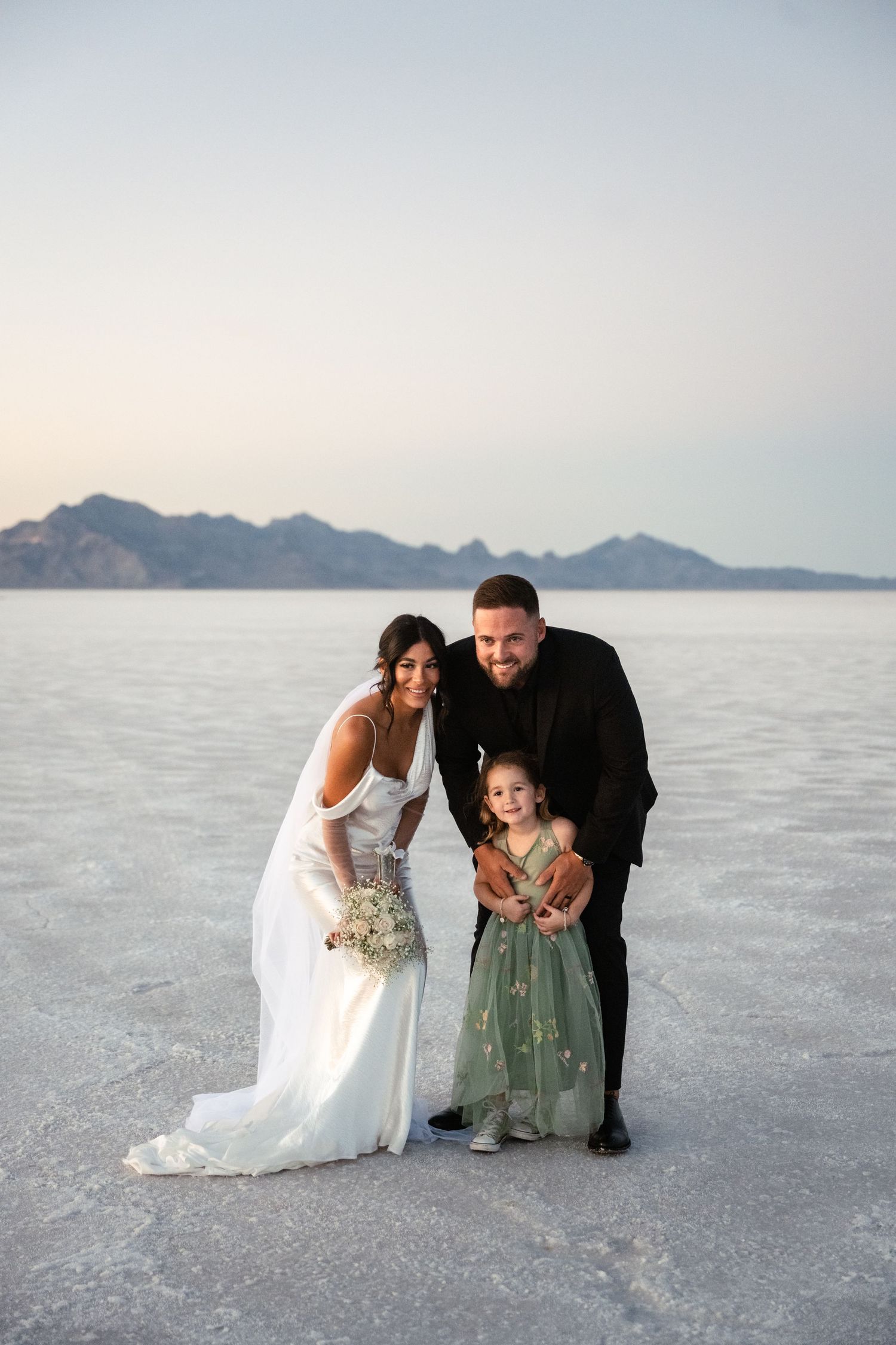 Wedding couple poses with child on salt flats with mountains in background.