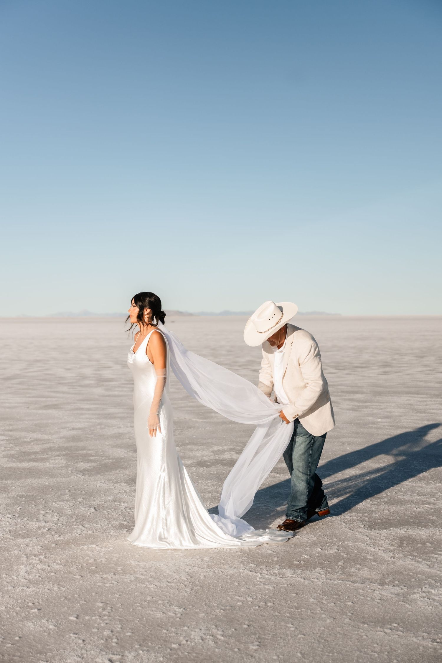 Wedding couple poses dramatically on salt flats with flowing veil in bright natural light.