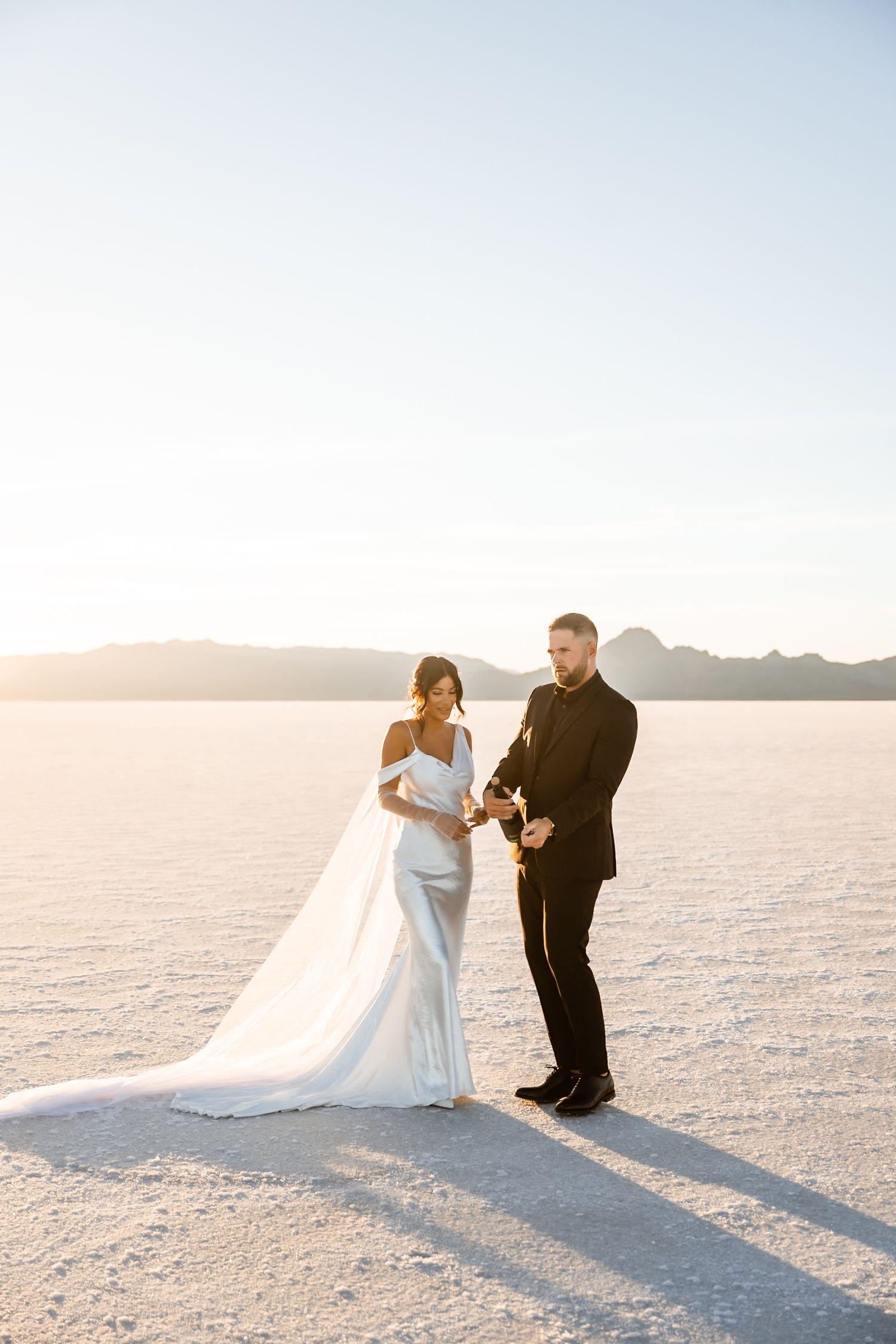 Wedding couple poses dramatically on white salt flats at sunset with mountains in background.