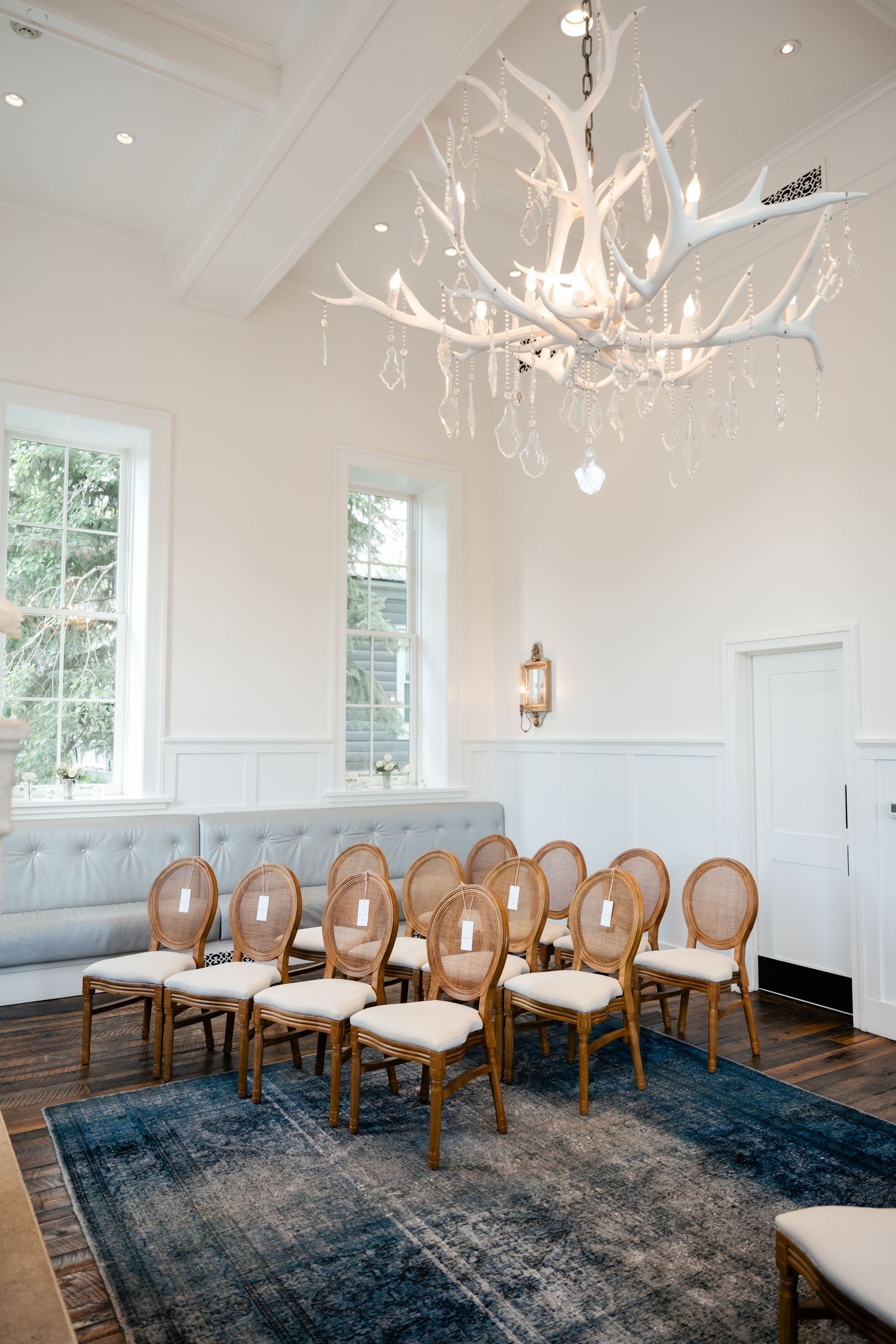 Elegant white antler chandelier hangs above wooden chairs with upholstered seats on a blue vintage rug in a bright room.