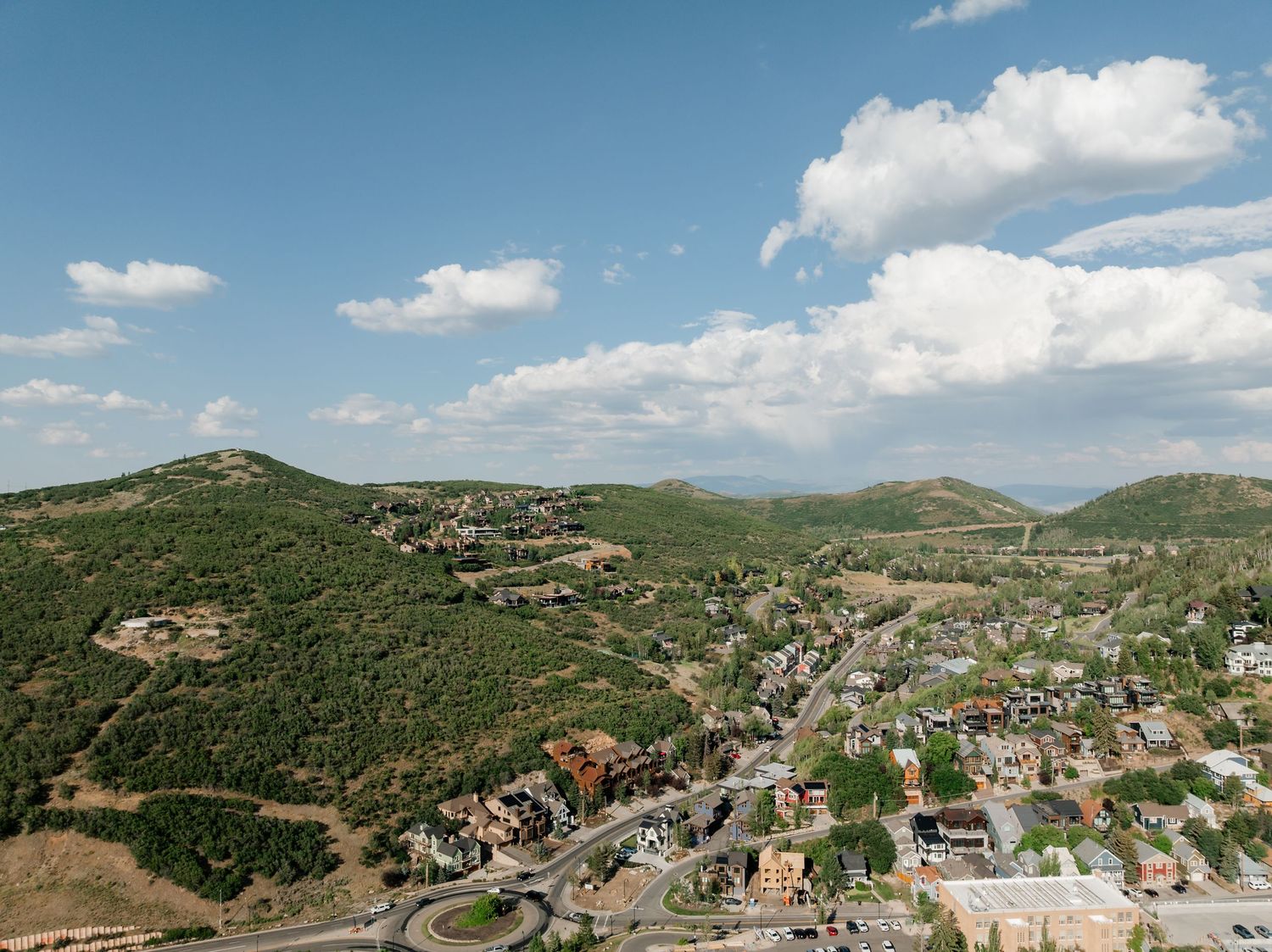 Panoramic aerial view of a small mountain town nestled in rolling green hills under blue skies with white clouds.