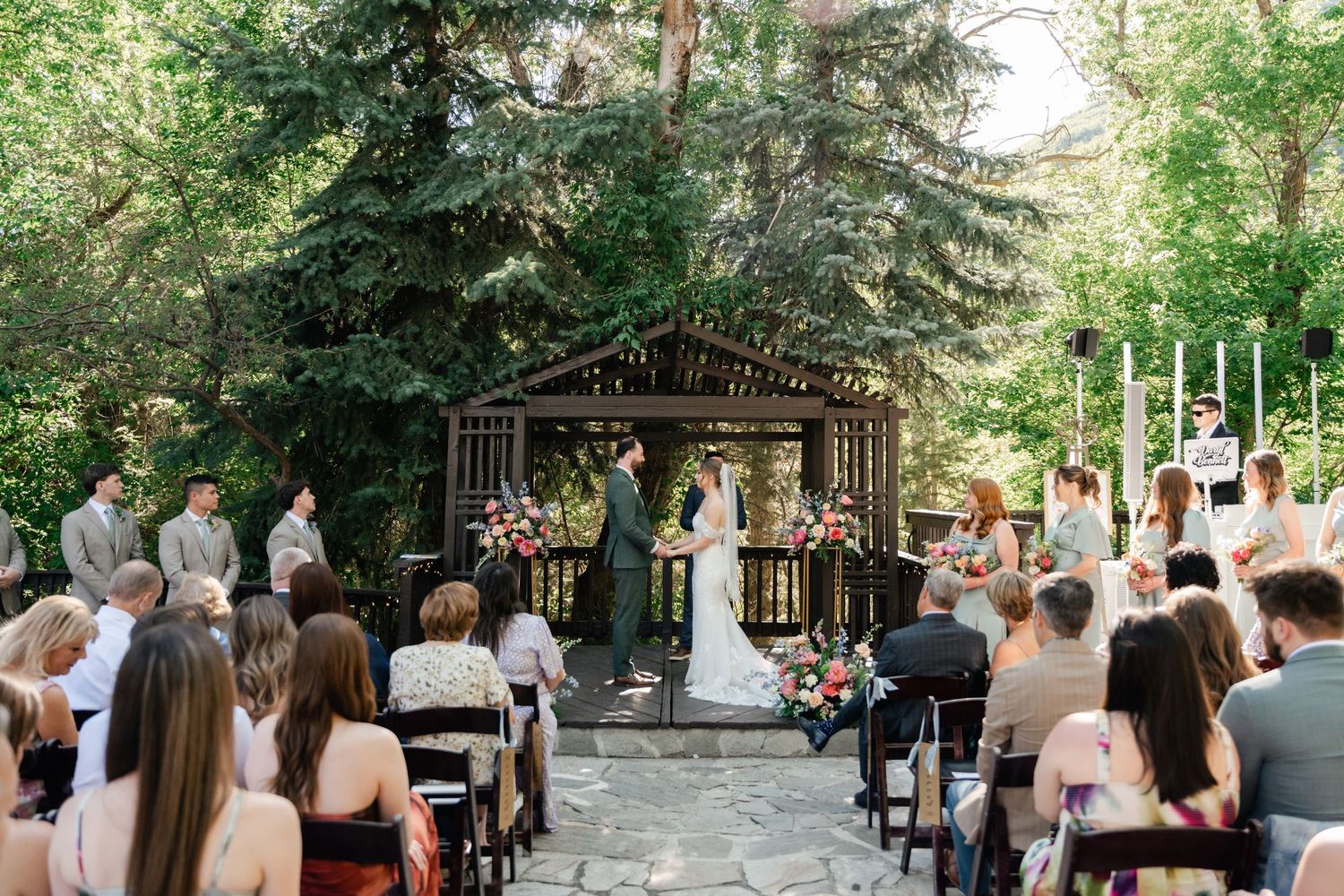 Wedding guests seated outdoors in white chairs watch a ceremony beneath tall evergreen trees in a forest setting. Millcreek Inn Wedding.