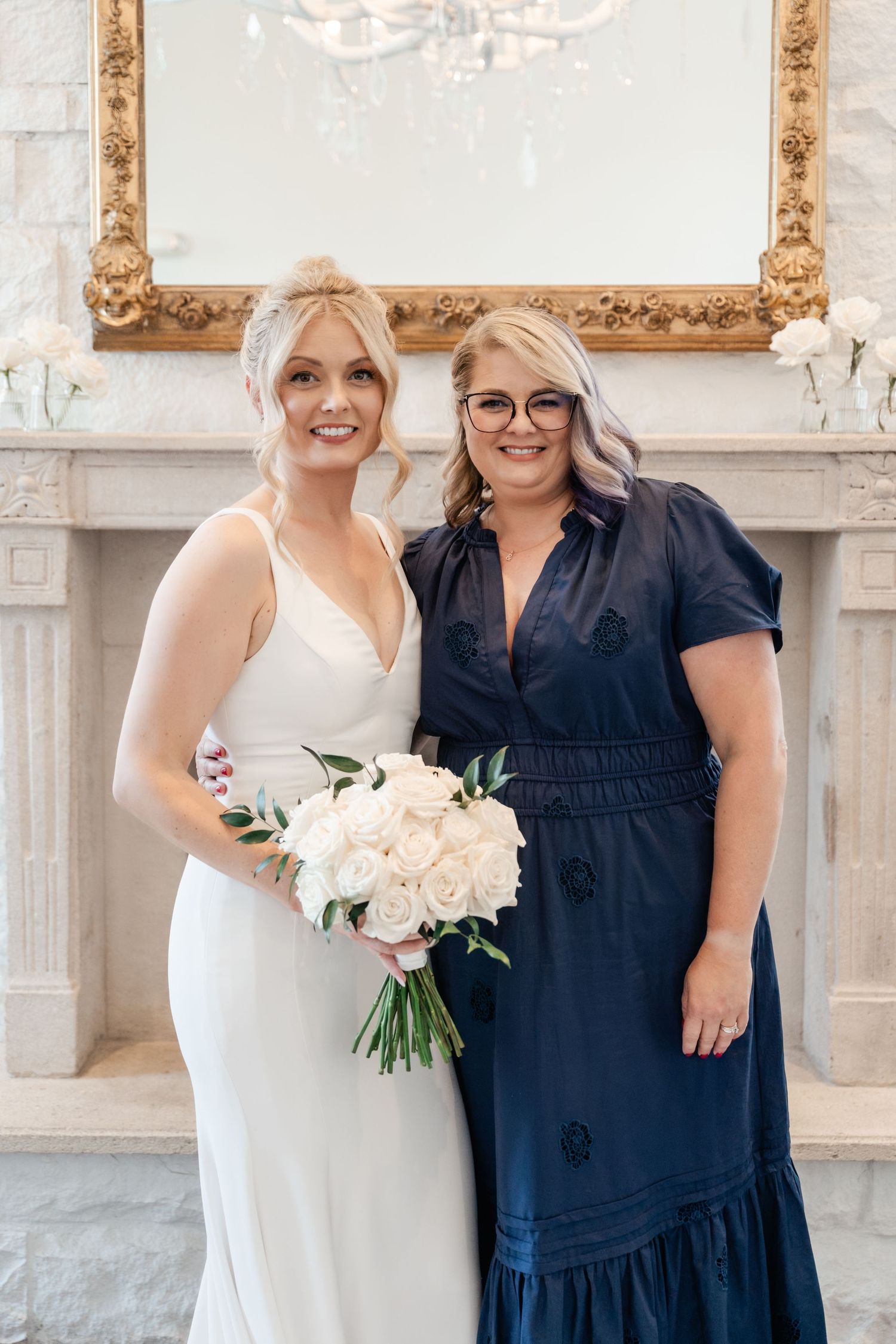 A bride in a white dress poses with a woman in navy blue holding white floral bouquets at an elegant venue.