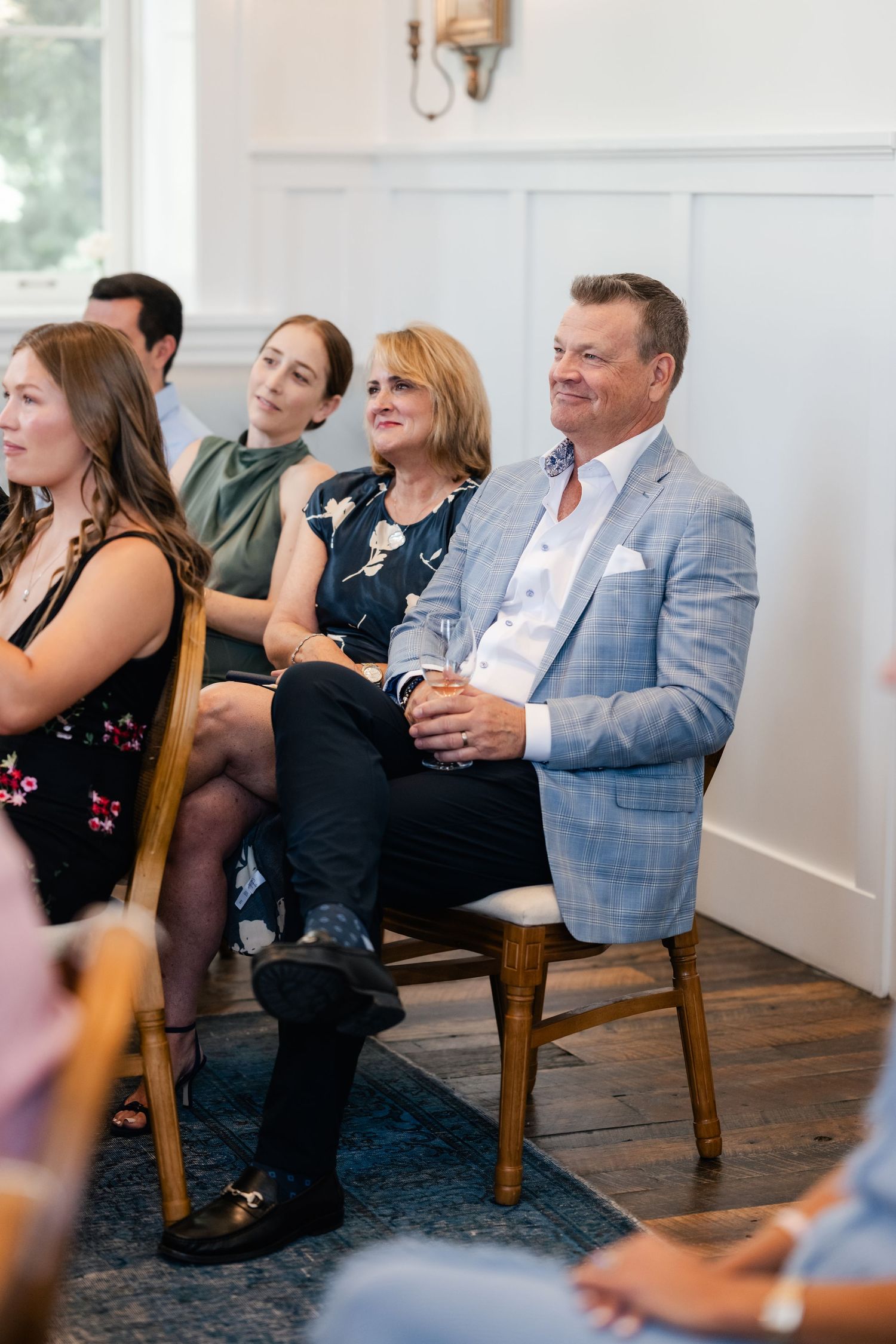 Wedding guests enjoy a lighthearted moment while seated in wooden chairs against white wainscoting.