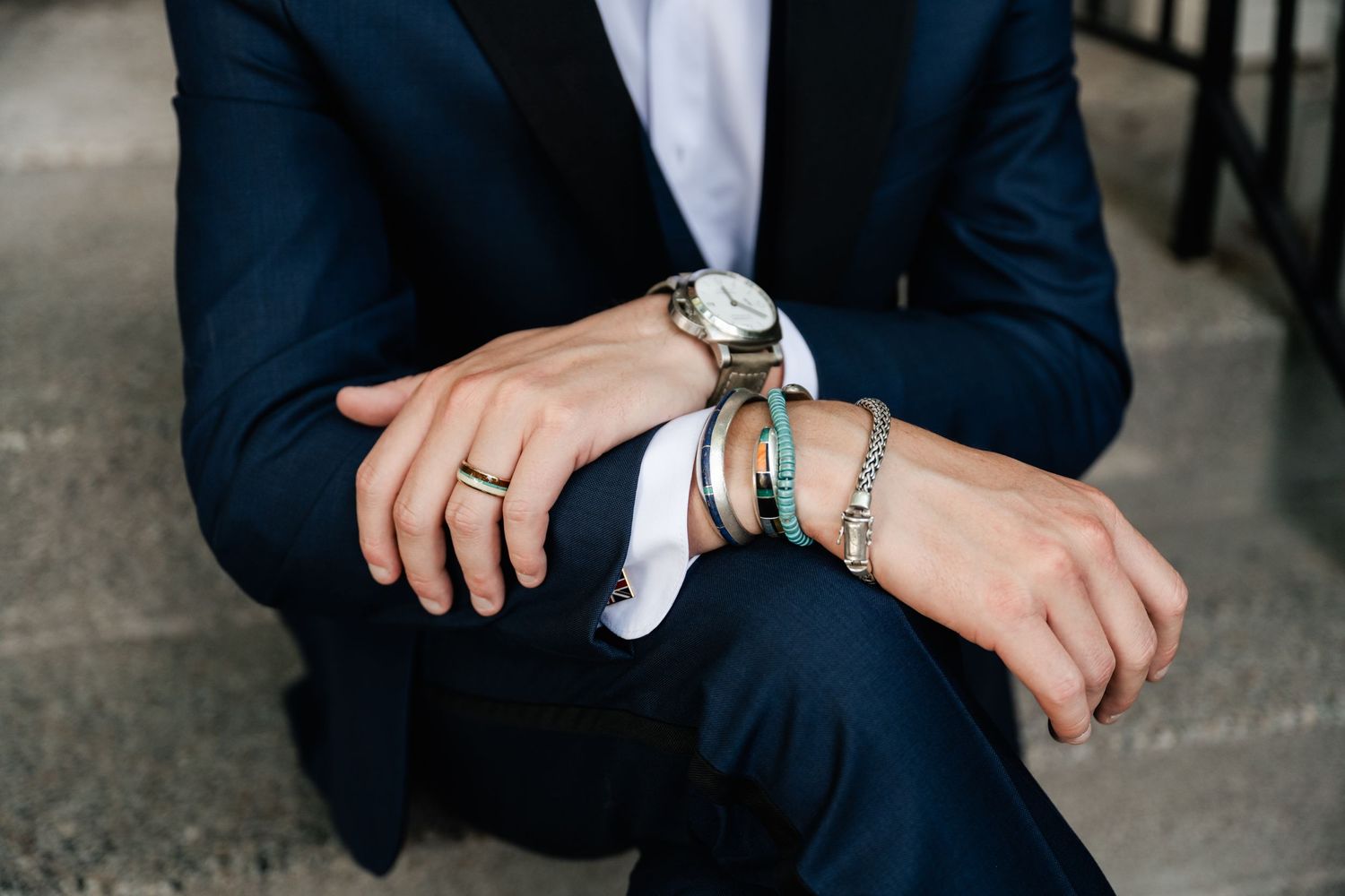 Close-up of hands wearing a navy suit sleeve showing silver watch, wedding ring and bracelet accessories.