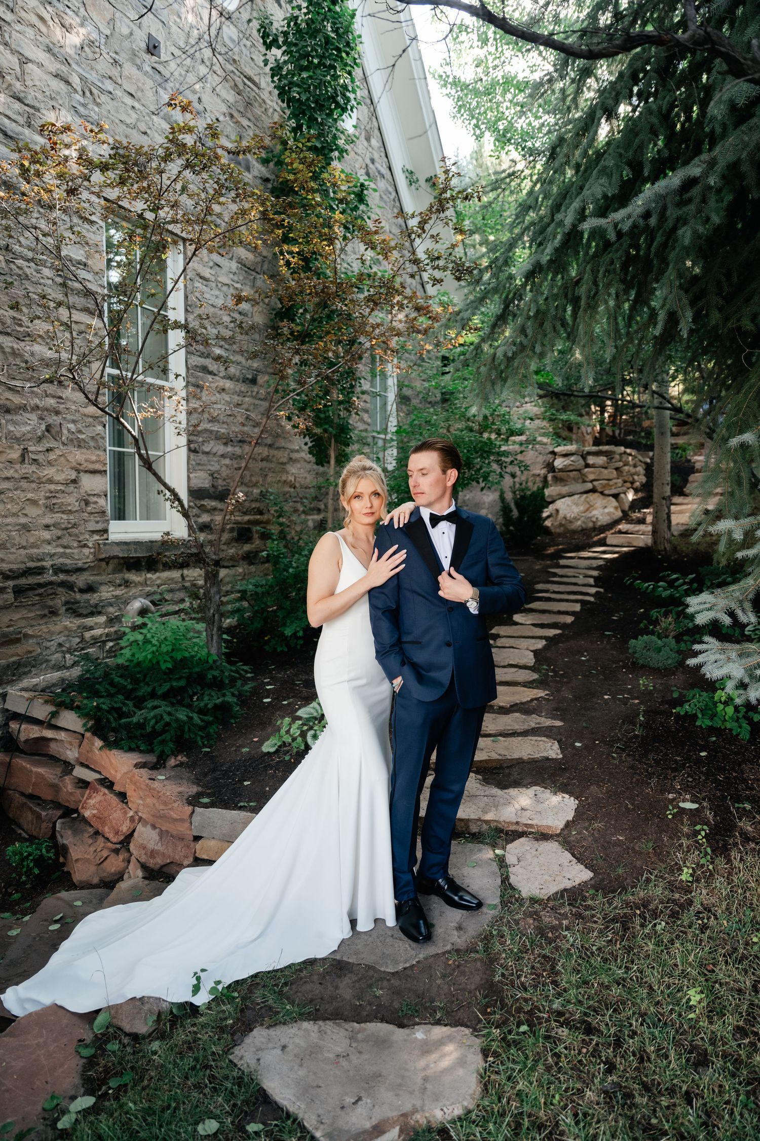 Wedding couple poses along a stone pathway surrounded by evergreen trees and natural landscaping.