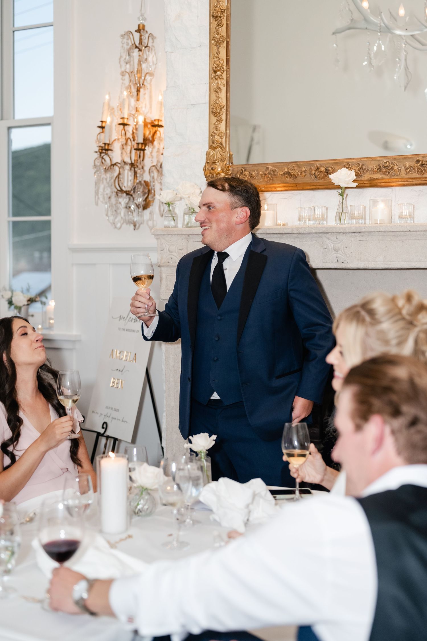 Guest making a toast at elegant wedding reception with crystal chandeliers and gold-framed mirror.