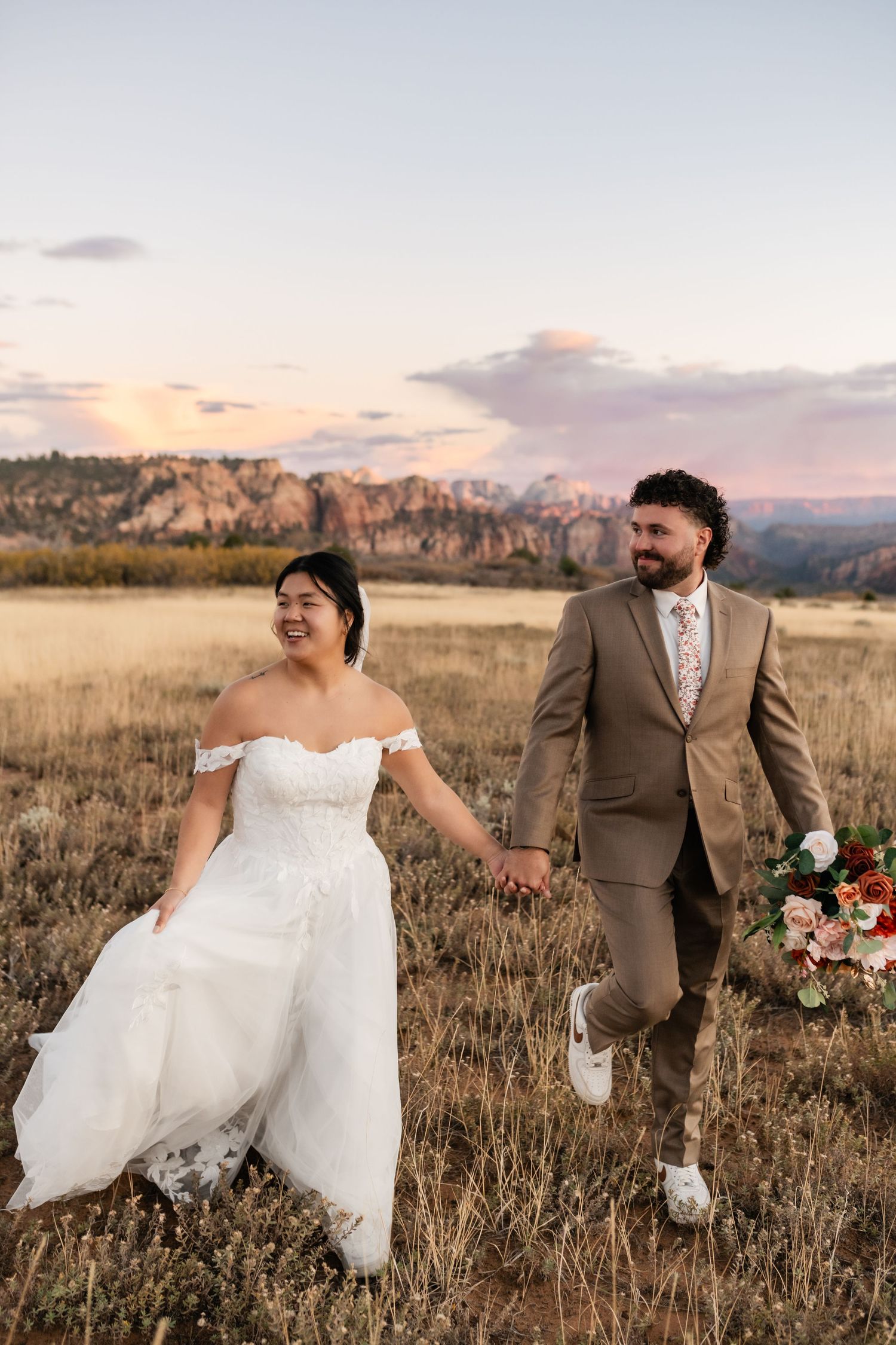 Wedding couple walking through a golden autumn field with mountains in the background at sunset.