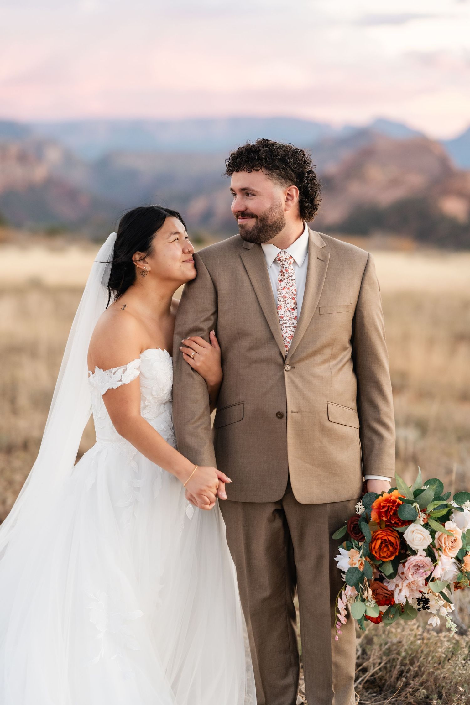 A wedding couple poses with a colorful bouquet in a field against mountain backdrop.