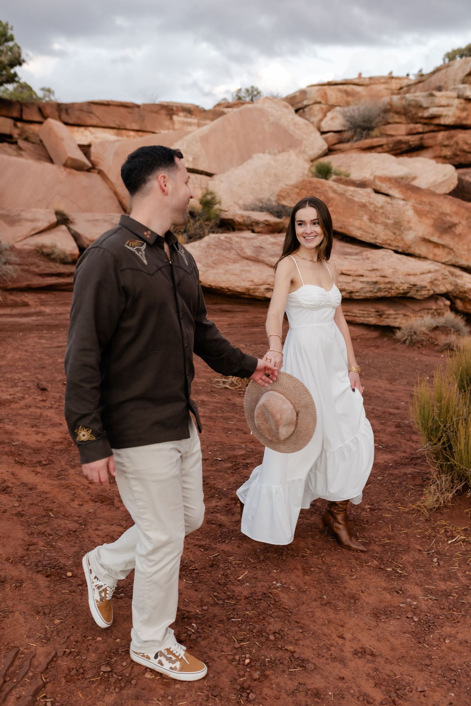 A couple walks together on red desert rocks while holding a brown hat.