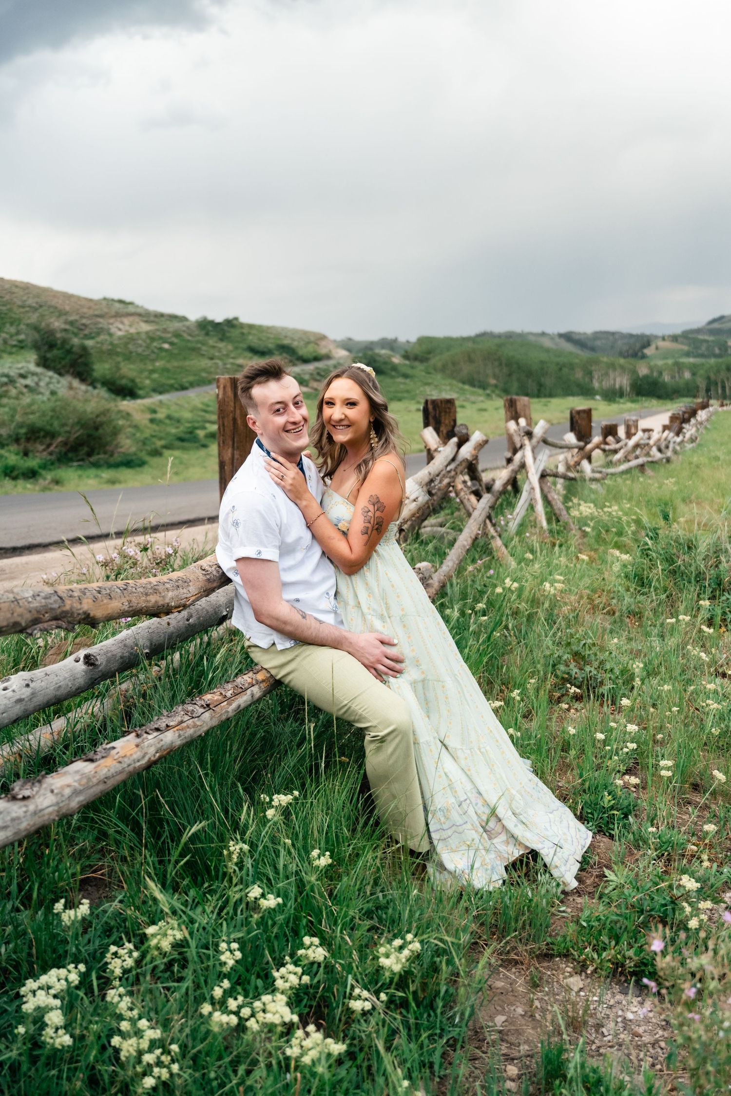Romantic couple shares joyful moments along a rustic wooden fence surrounded by wildflowers in a mountain meadow.