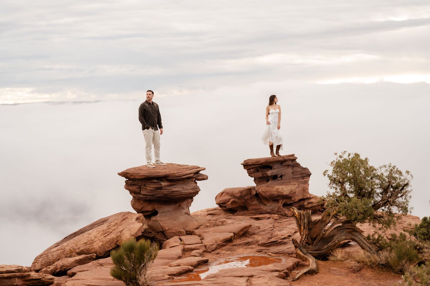 Silhouetted figures stand atop red rock formations against a cloudy sky at sunset in the desert landscape.