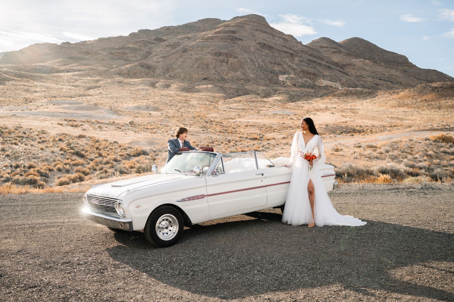 A classic white convertible in a desert landscape with a wedding couple with mountains and blue sky in the background. Utah Wedding.