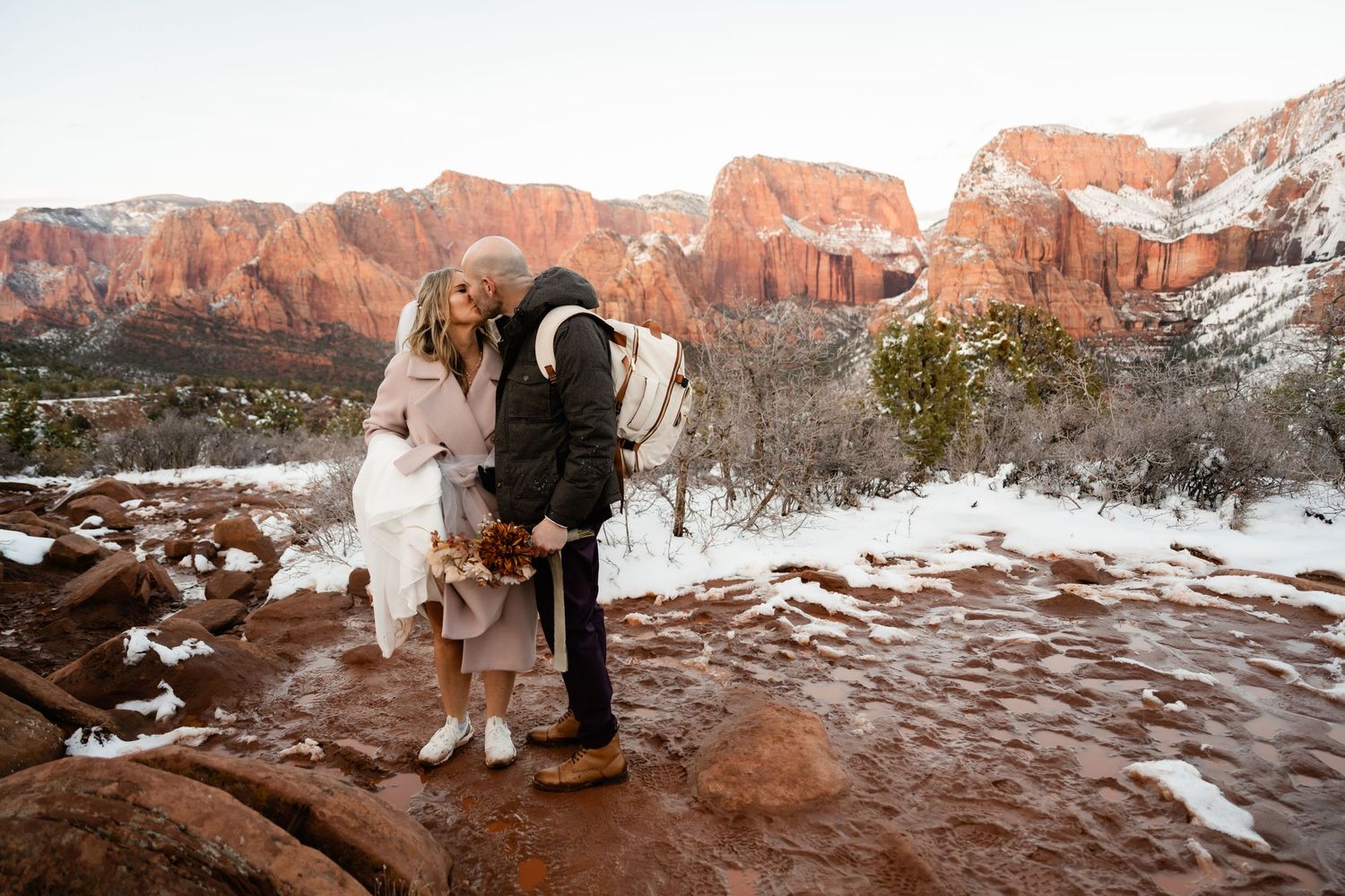 Wedding couple walking along snowy trail with towering red rock formations in background.