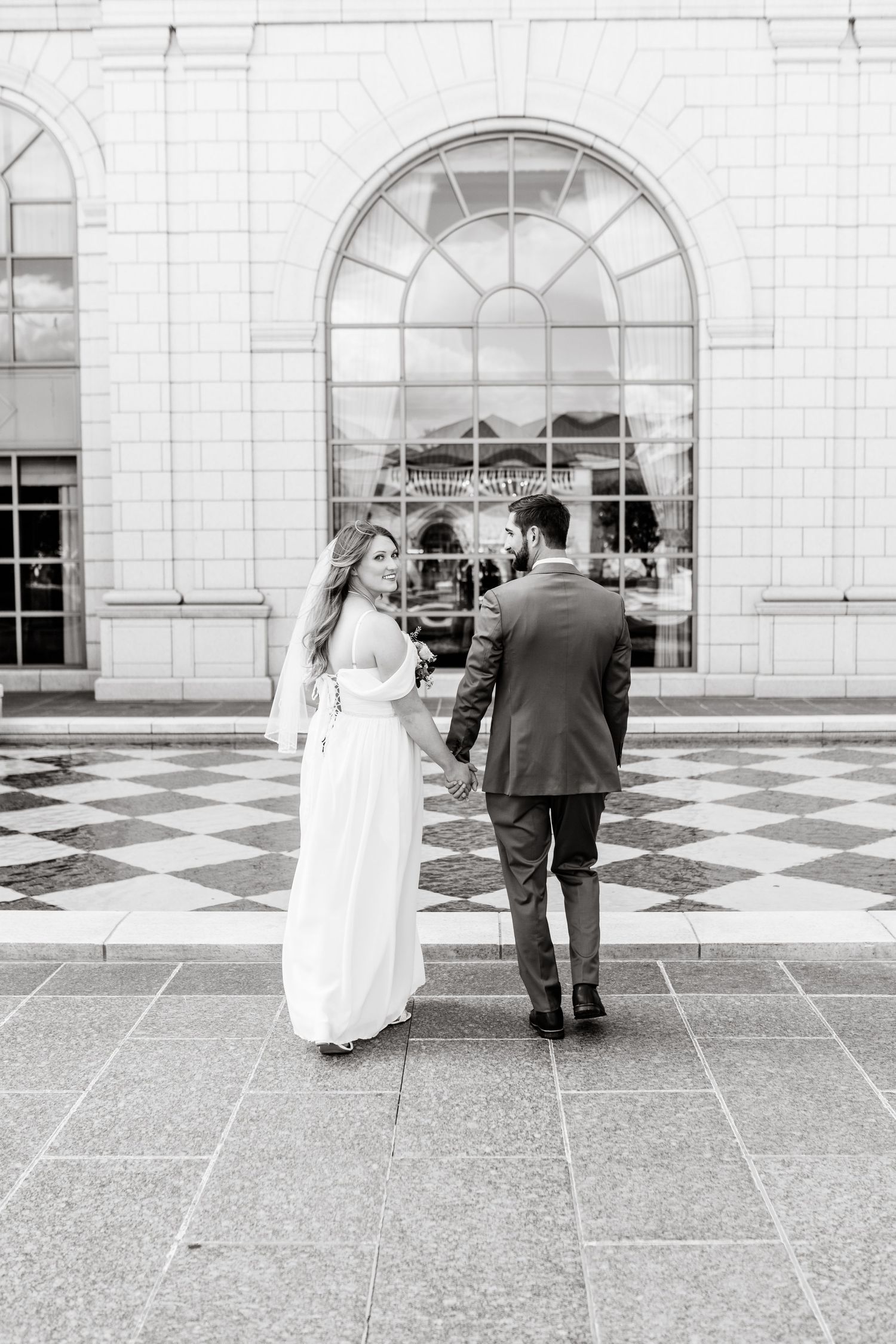 Black and white photo of a couple in wedding attire walking together on a checkered courtyard with arched architecture.