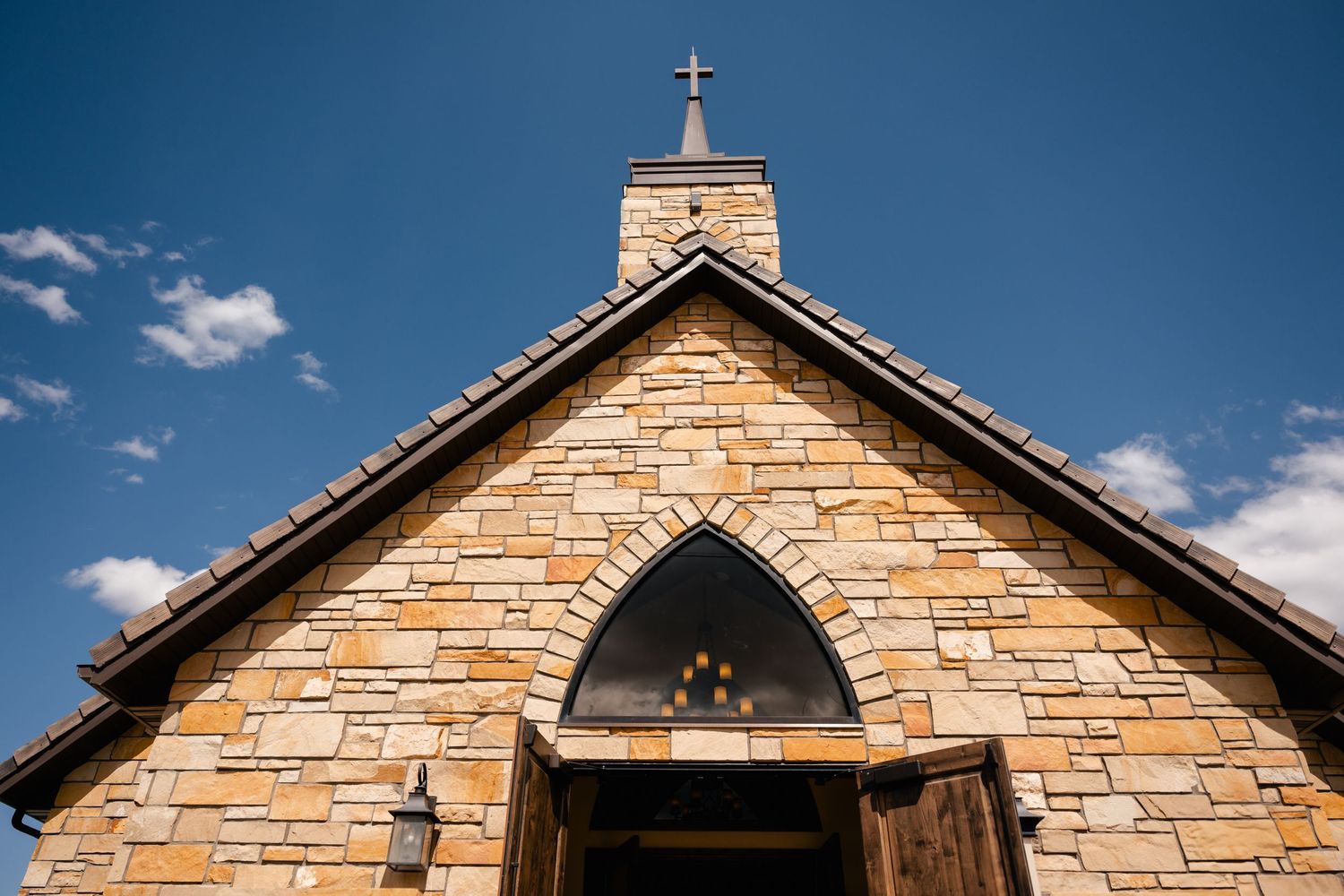 Stone chapel exterior with cross on top against blue sky with white clouds.