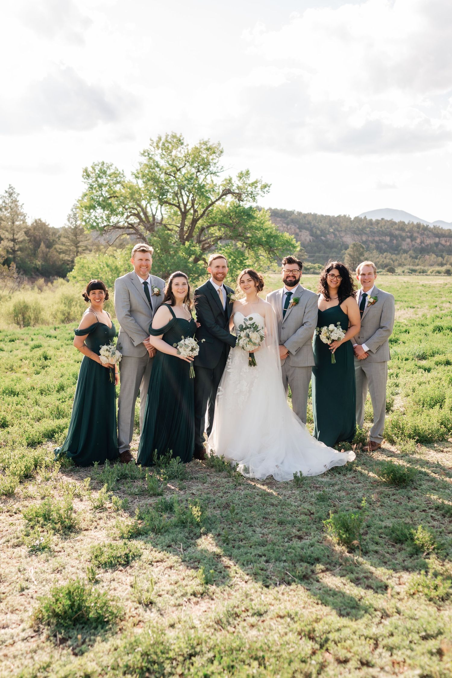 Wedding party in emerald green dresses and gray suits poses in natural mountain setting.