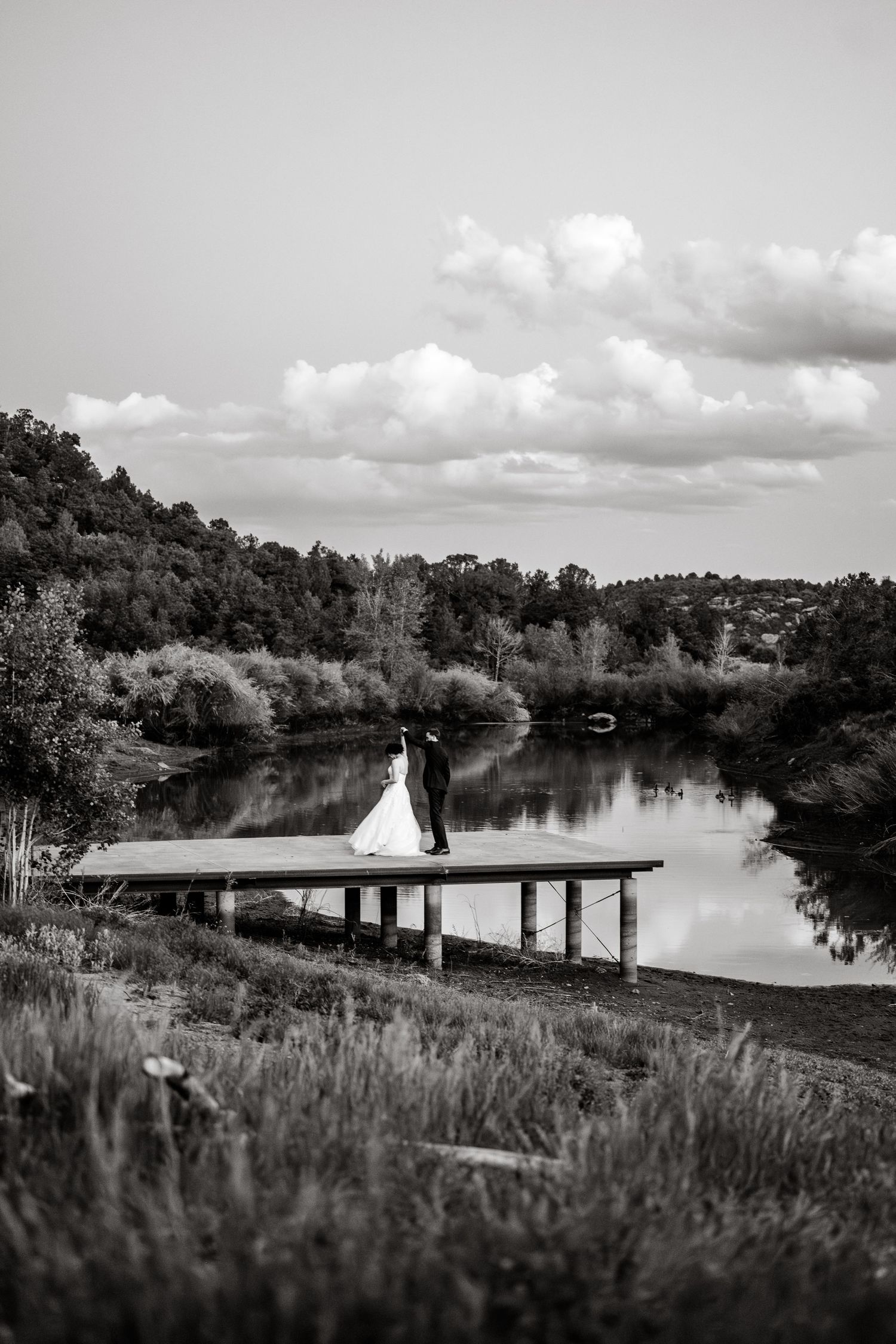 A scenic black and white panorama of a wooden dock extending over a tranquil lake surrounded by trees.