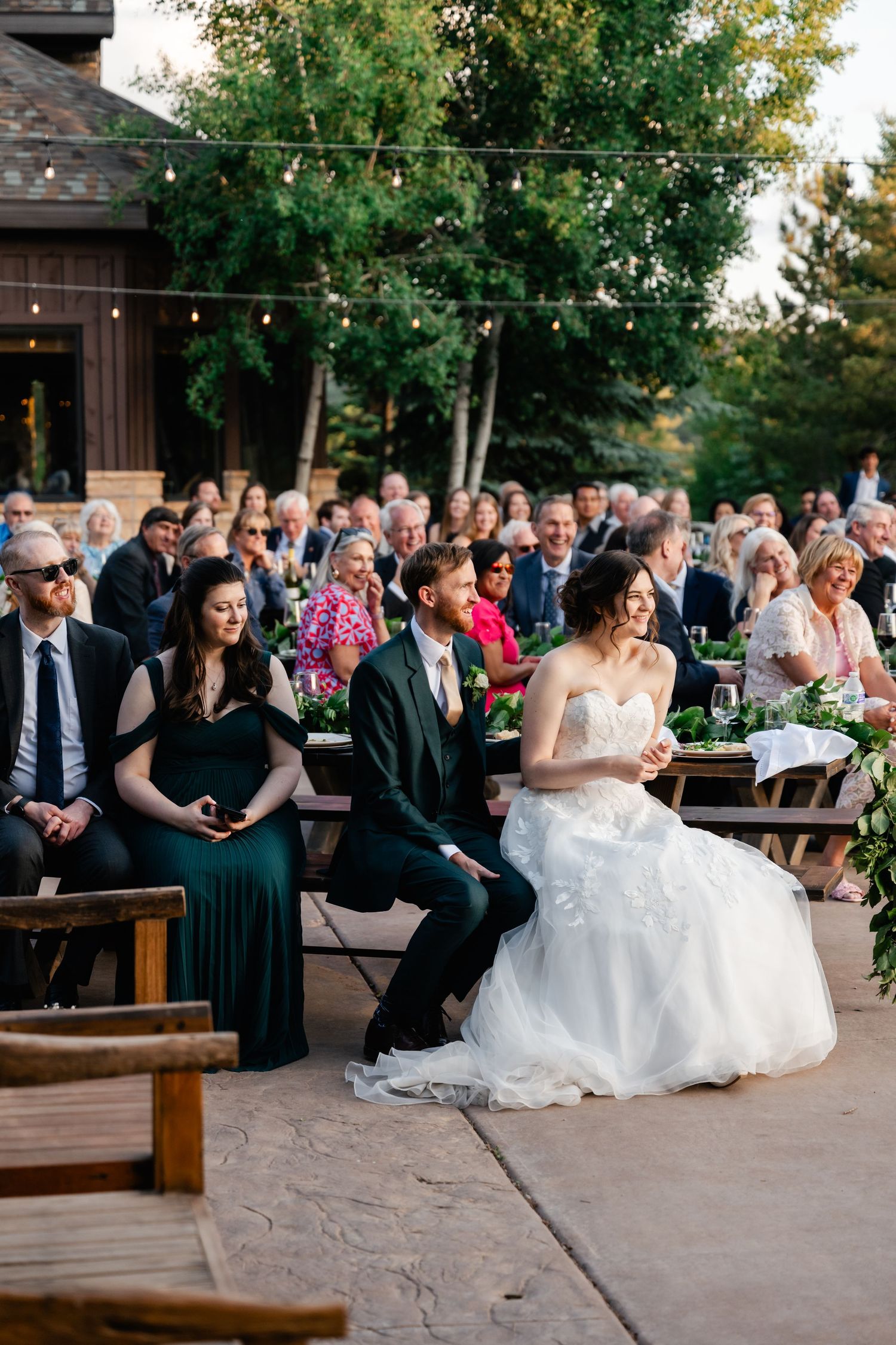 Outdoor wedding ceremony seating area with guests gathered on benches under string lights and trees.