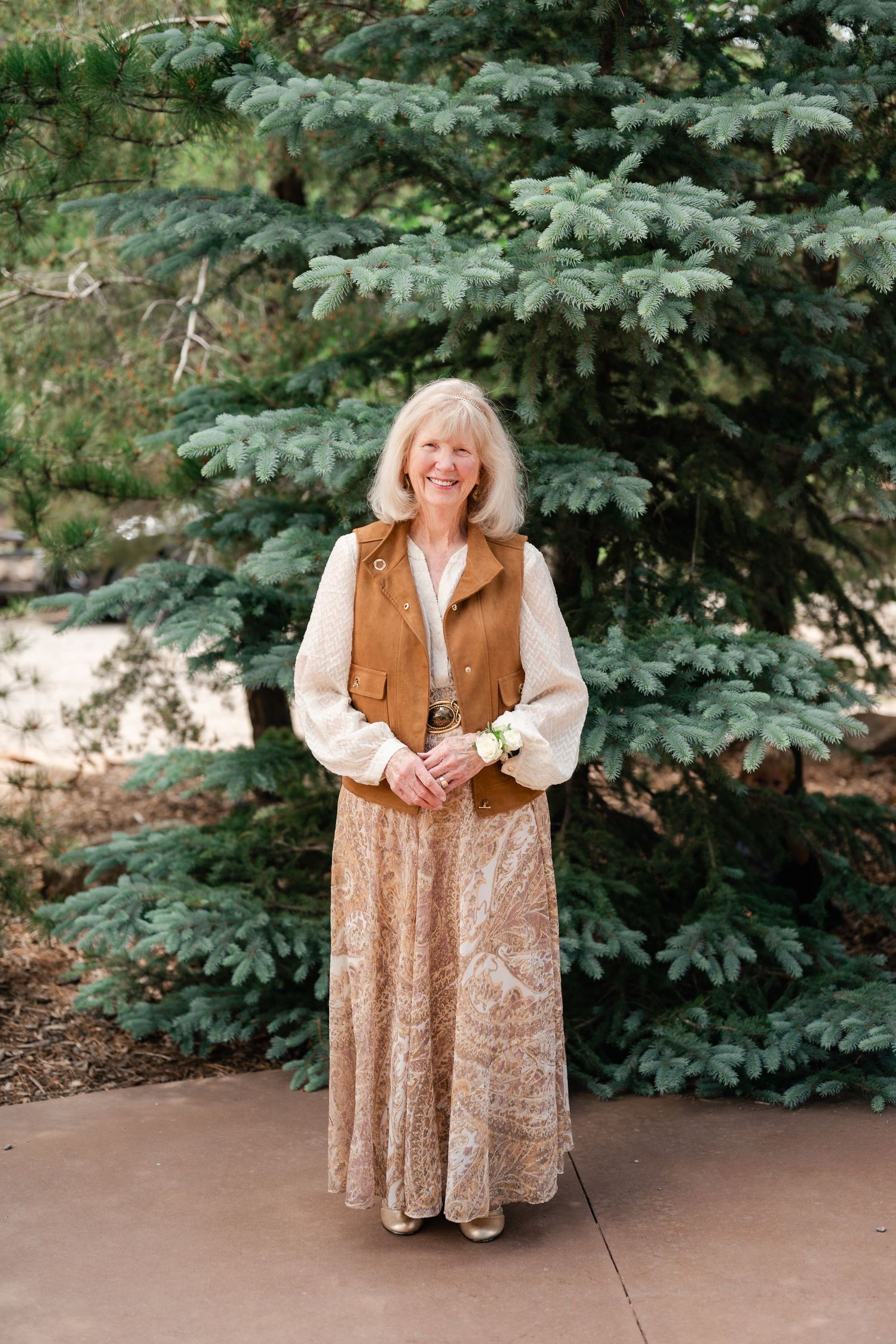 A person in a brown vest and floral dress poses outdoors against a backdrop of evergreen trees.