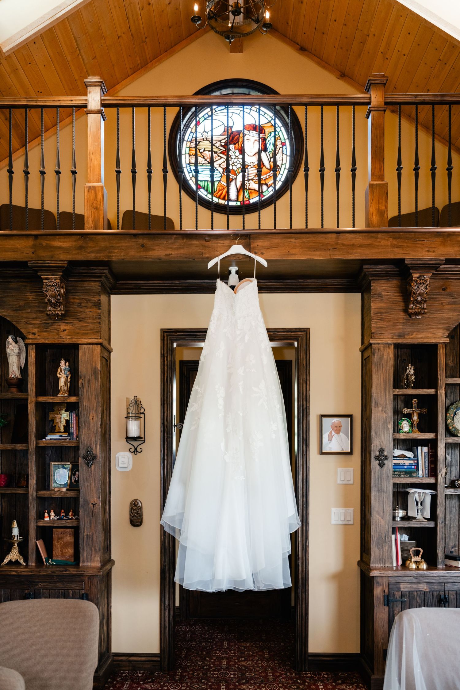 White wedding dress hanging in rustic venue interior with stained glass windows and wooden beams.