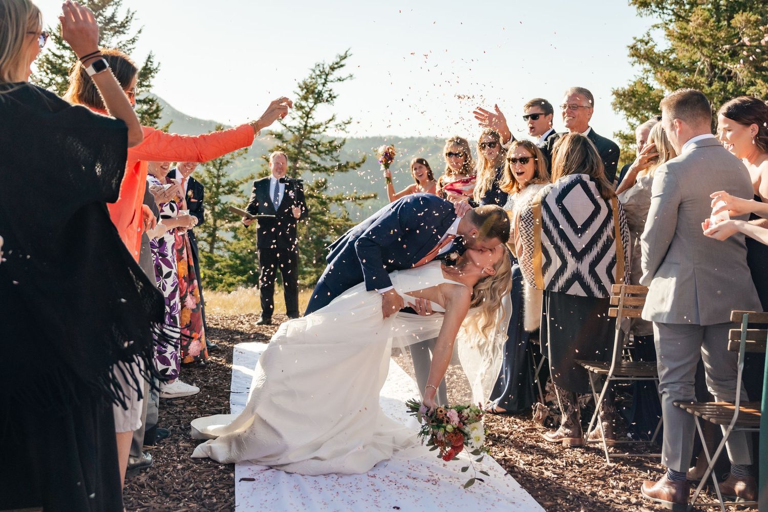 Wedding guests celebrate and throw confetti during an outdoor mountain ceremony.
