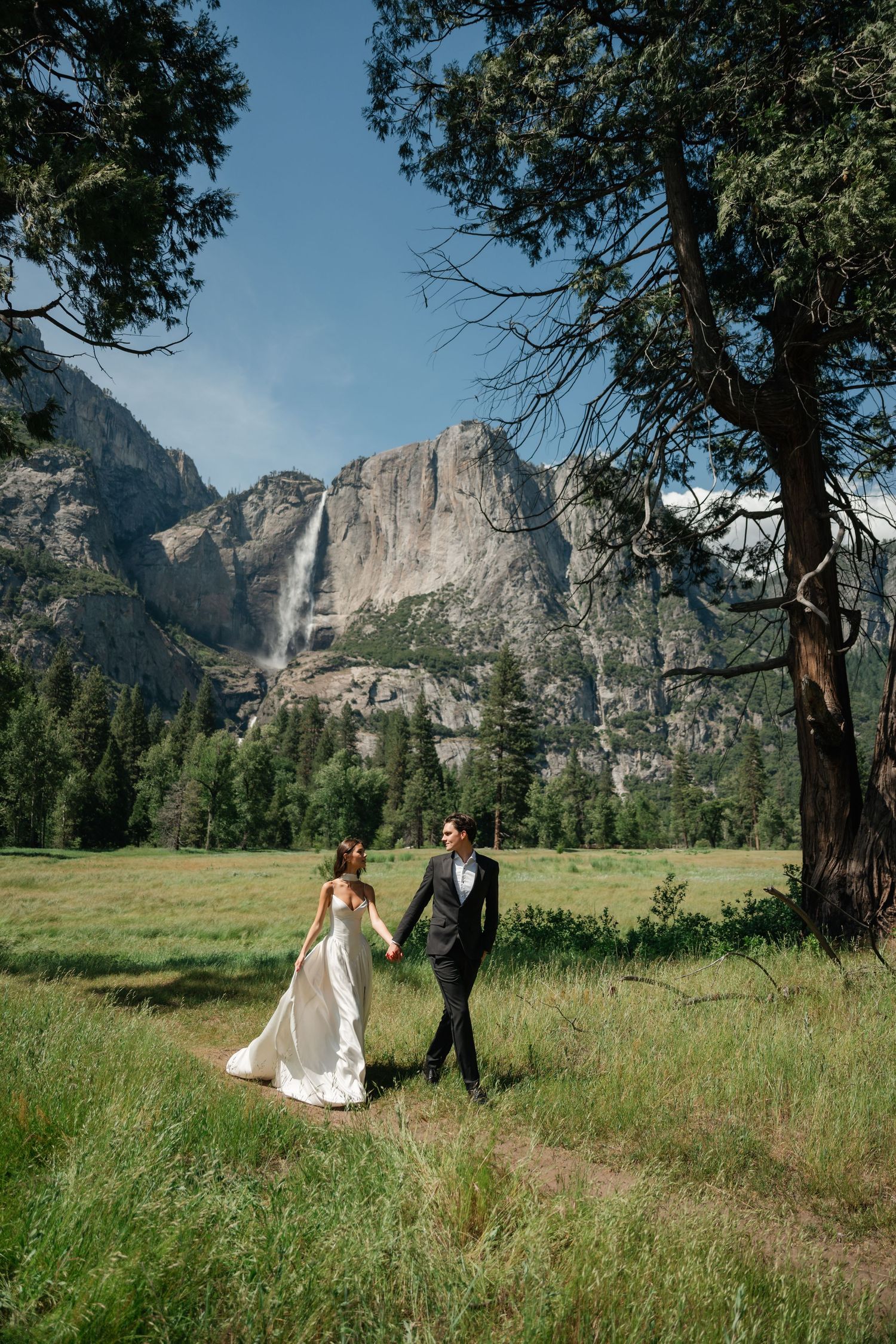 Bride and groom hold hands walking in Yosemite meadow with waterfall and granite cliffs backdrop.