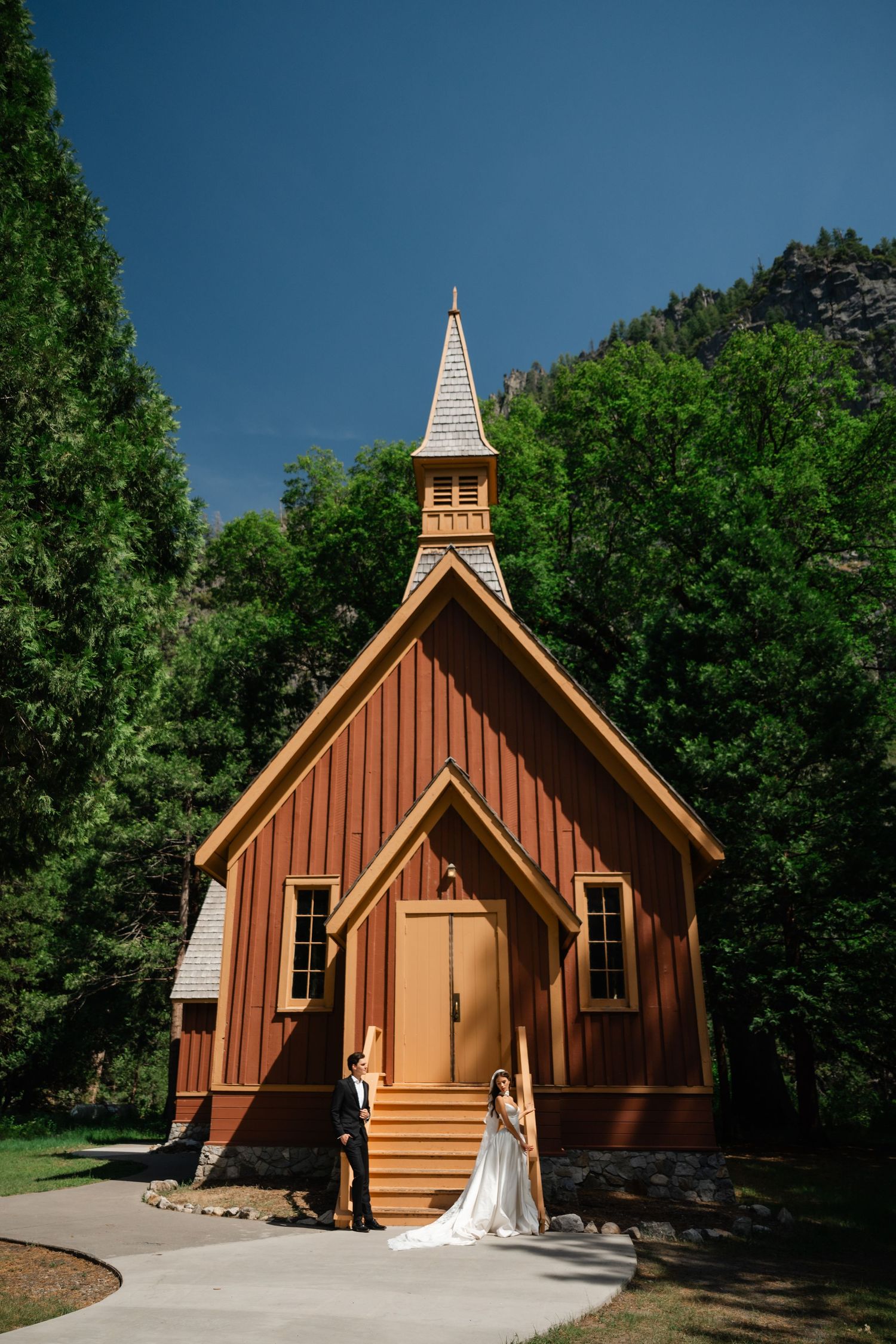 Bride and groom pose before a rustic red wooden chapel in Yosemite Valley, surrounded by lush green trees.
