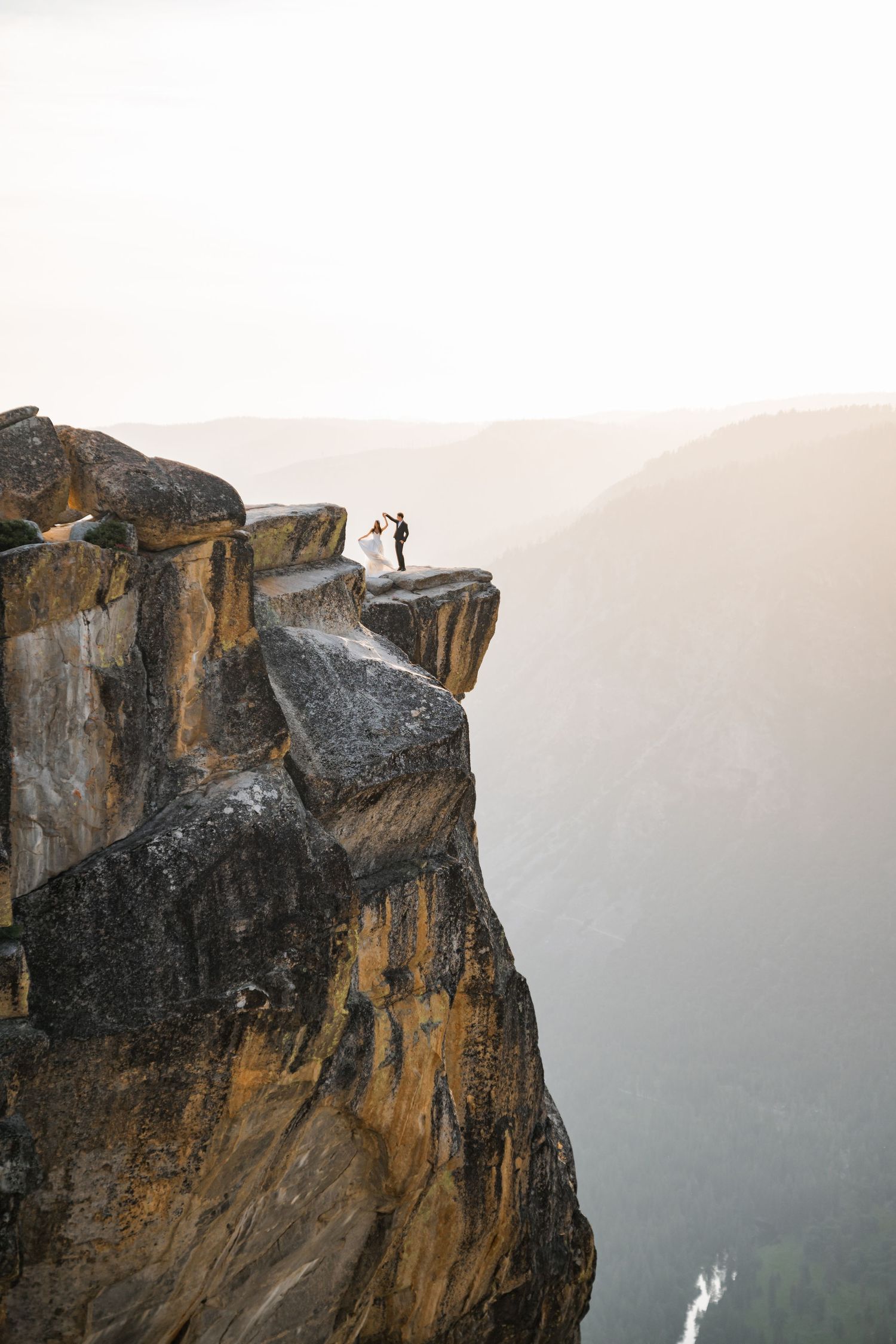Bride and groom stand on dramatic cliff edge overlooking vast mountain valley at golden hour.