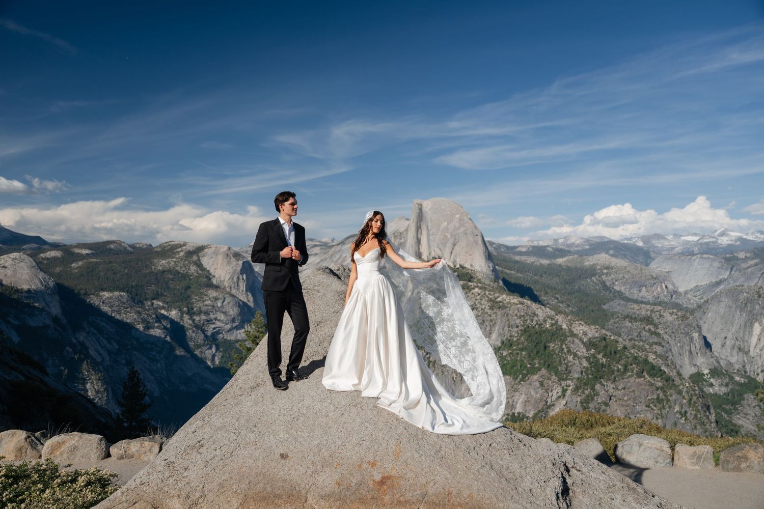 Bride and groom stand on rocky summit at Yosemite with Half Dome and blue skies behind them.