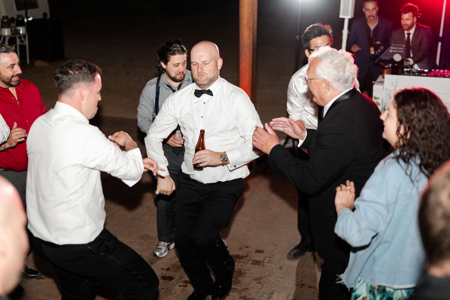 Men in formal attire dancing energetically at a wedding reception party indoors at night.