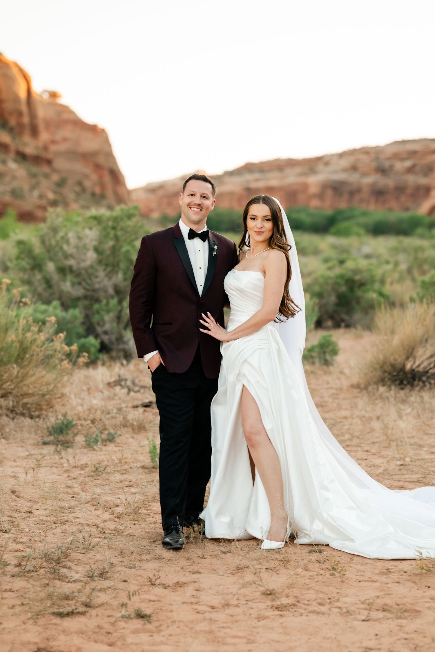 Bride and groom pose together in desert landscape with red rock formations at golden hour.