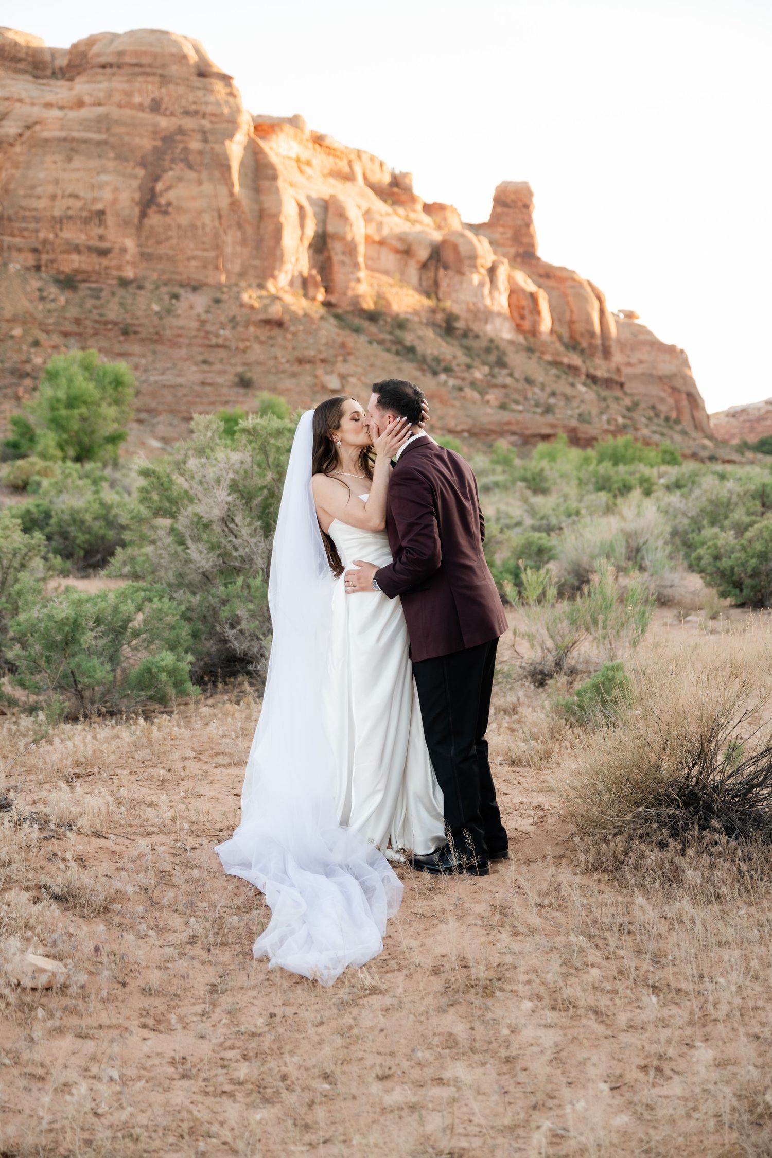 Bride and groom kissing in a desert landscape with red rock formations and green shrubs in the background.