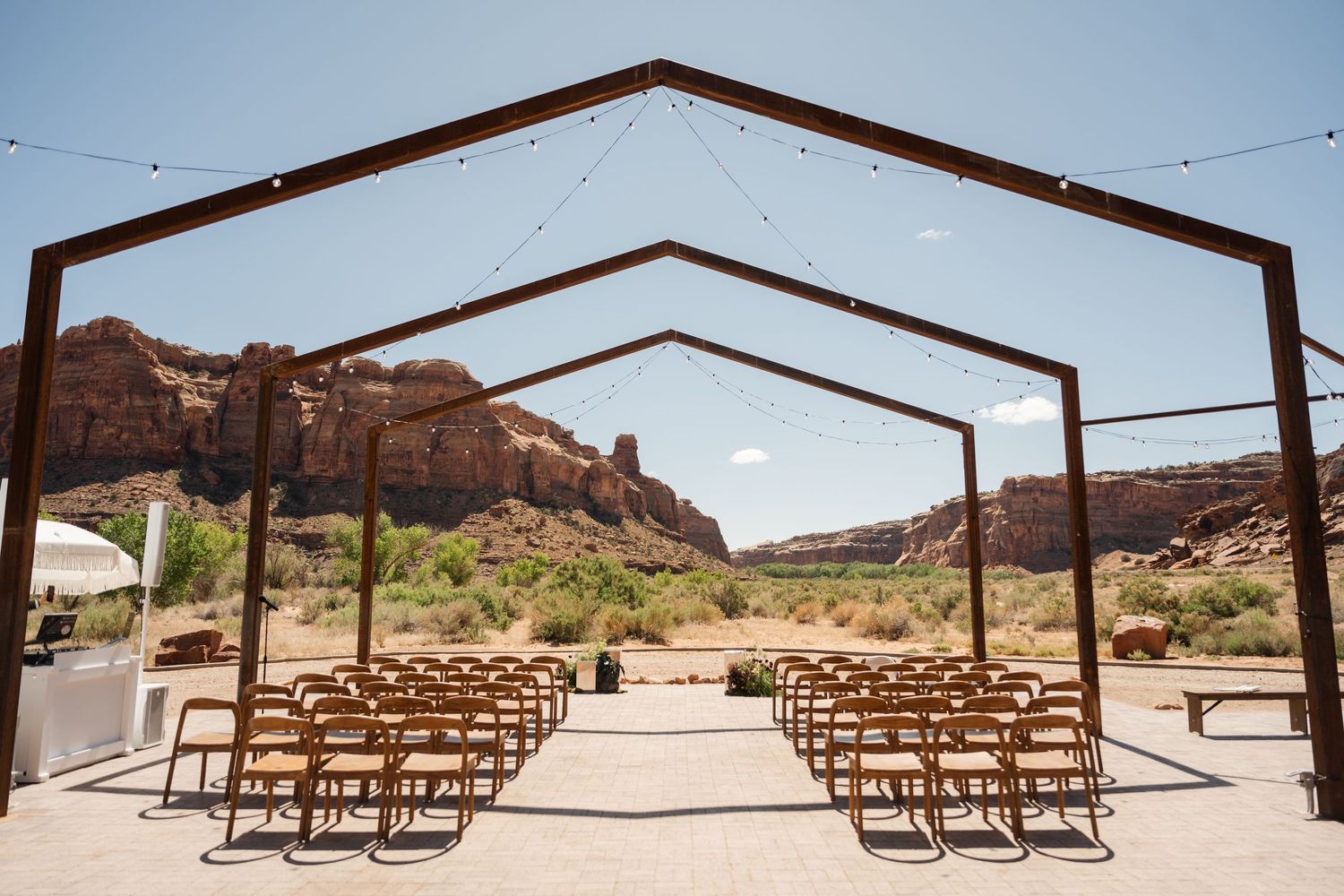The Red Earth Venue - Outdoor desert wedding ceremony venue with wooden chairs under a modern steel pergola, red rock canyon backdrop.