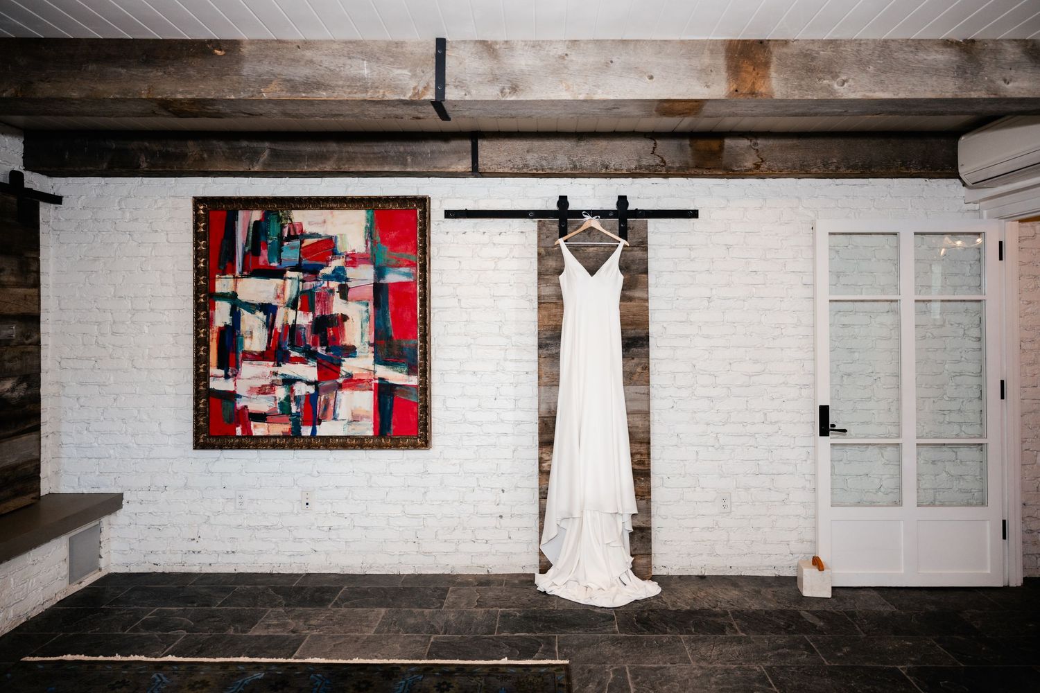 White wedding dress hanging on a barn-style rail against a white brick wall beside a vibrant abstract painting.