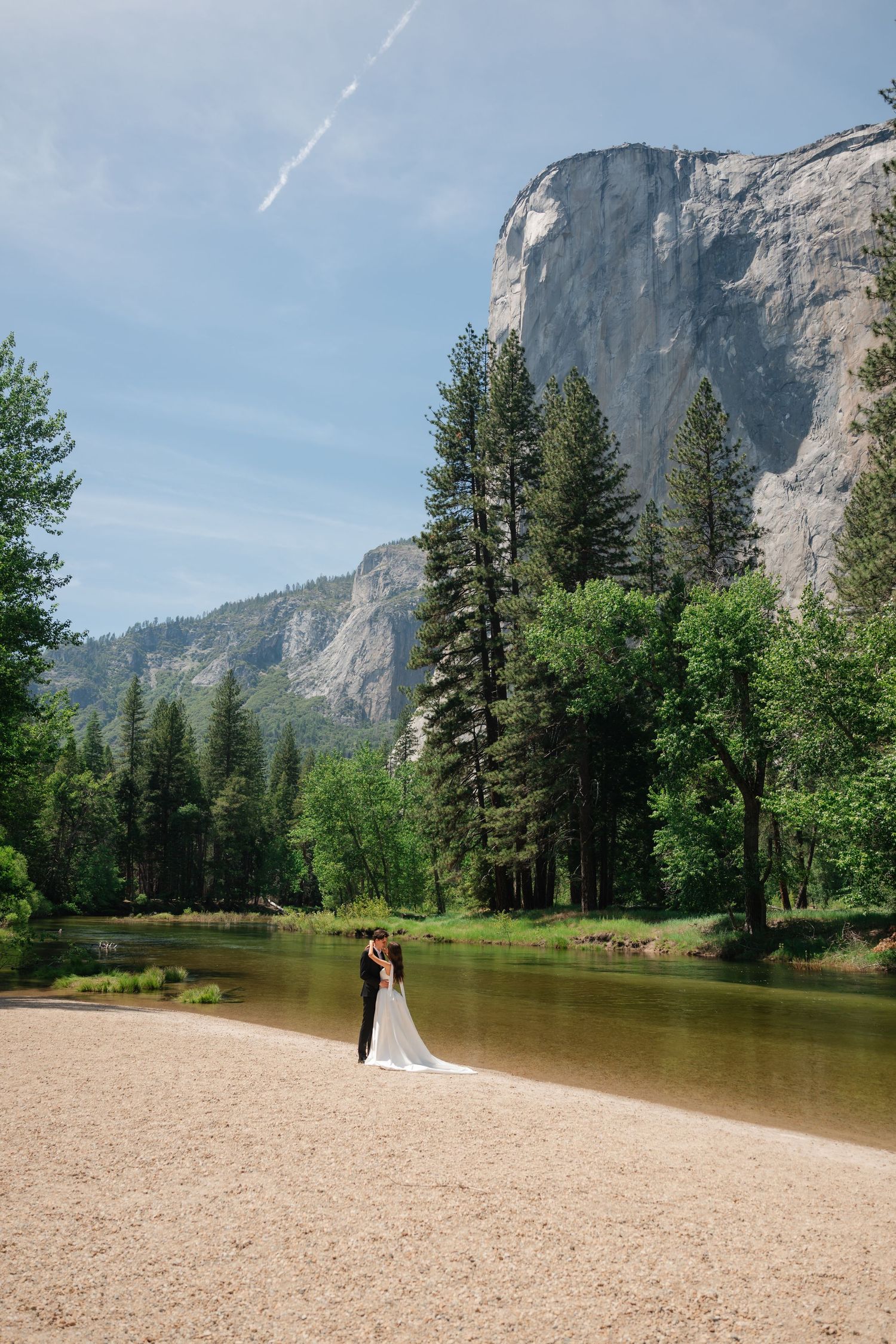 Wedding couple embraces on sandy riverbank with majestic Yosemite granite cliffs and tall pines backdrop.